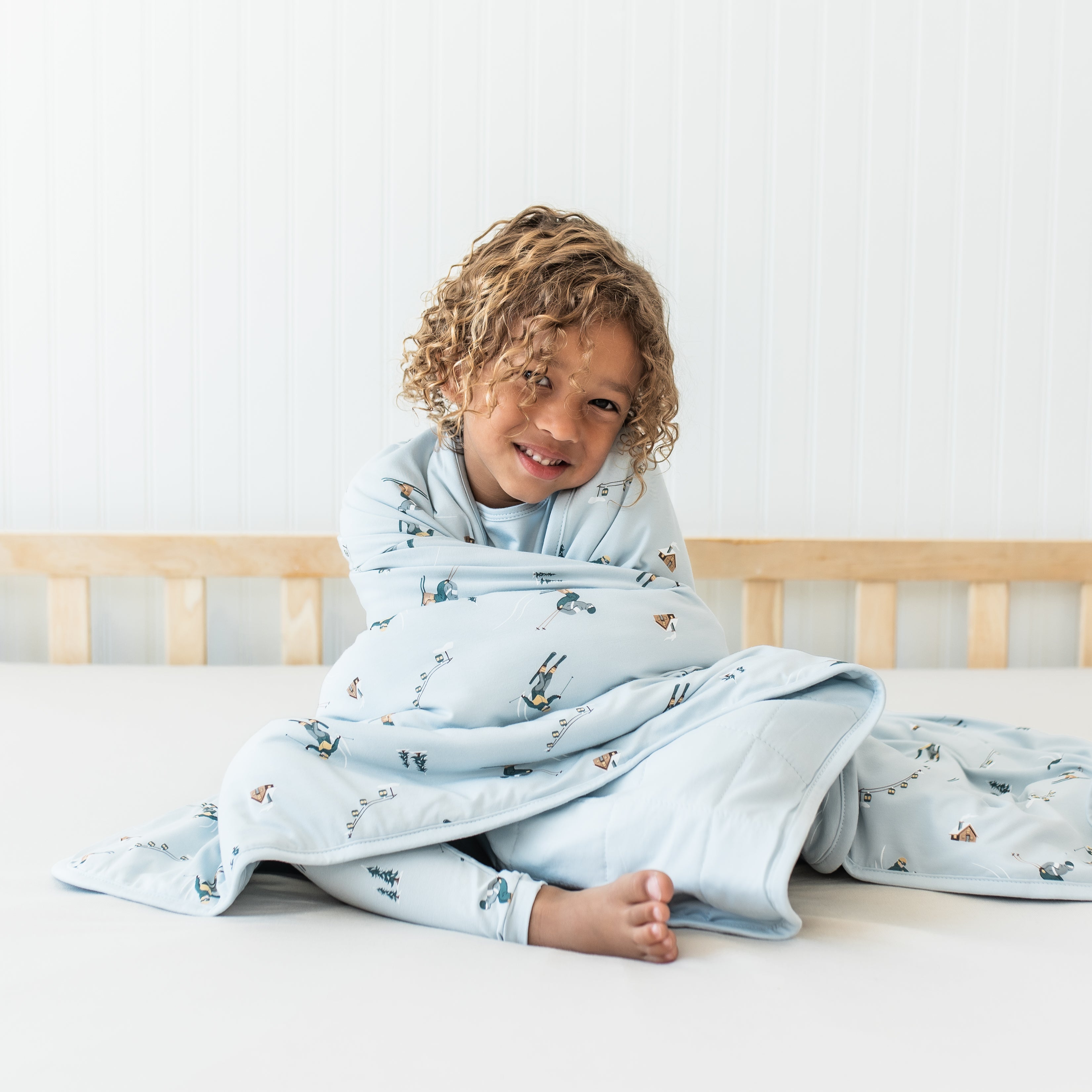 Young boy sitting on a bed with the Toddler Blanket in Ski 1.0 wrapped around him