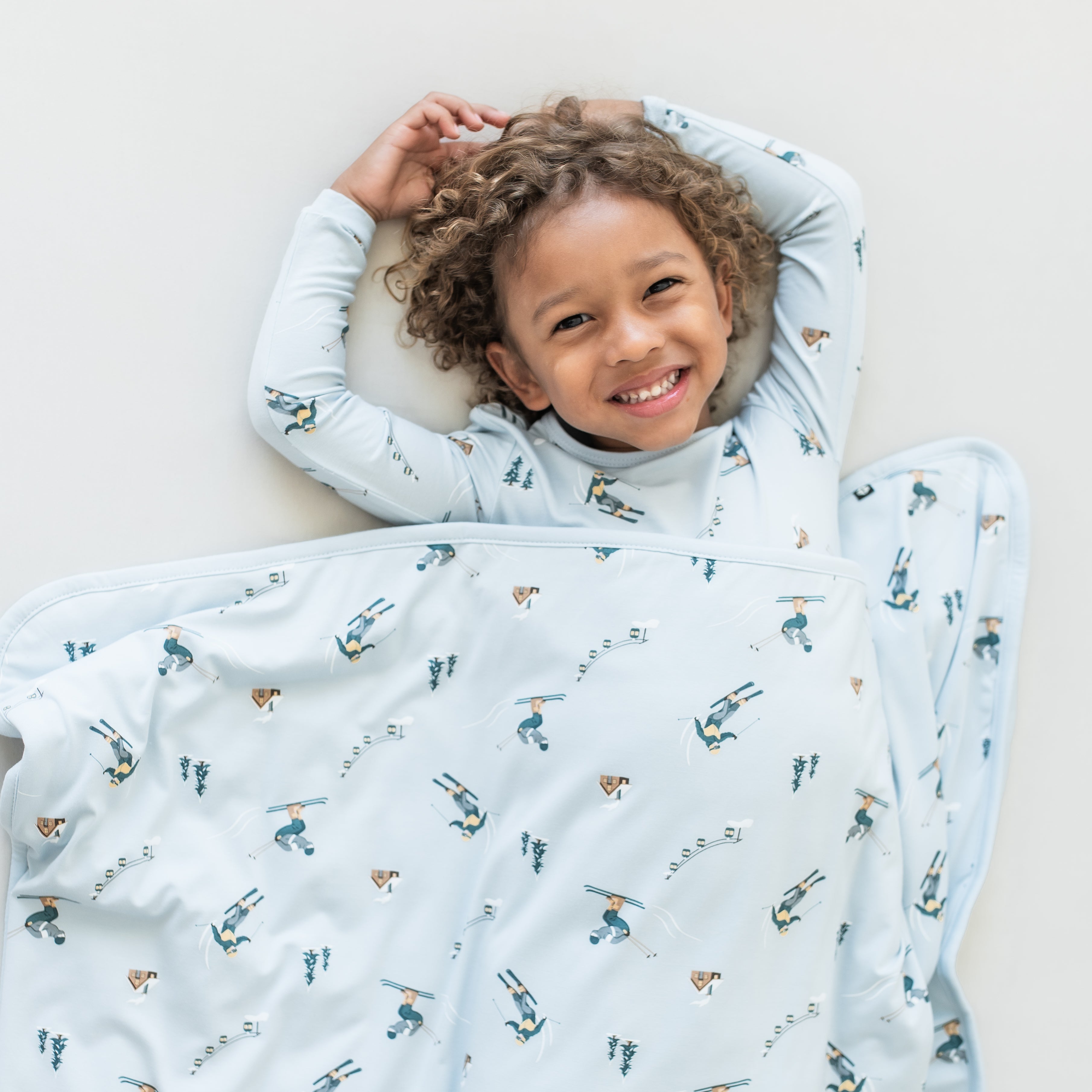 Smiling boy laying down on a light neutral surface with the Toddler Blanket in Ski 1.0 overtop of him