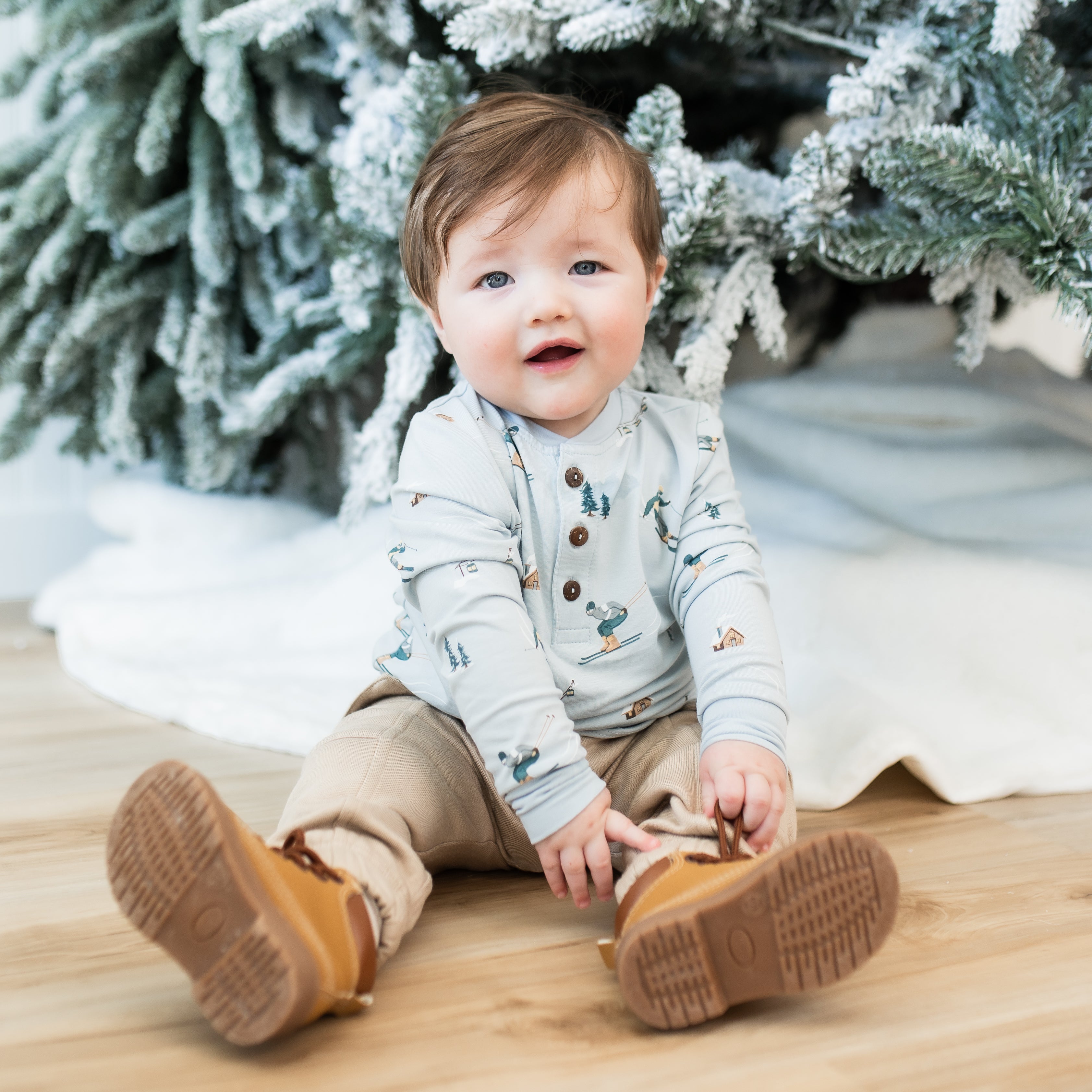 Young toddler sitting on a light wood floor in front of a frosted tree wearing the Long Sleeve Toddler Henley Tee in Ski with light tan pants