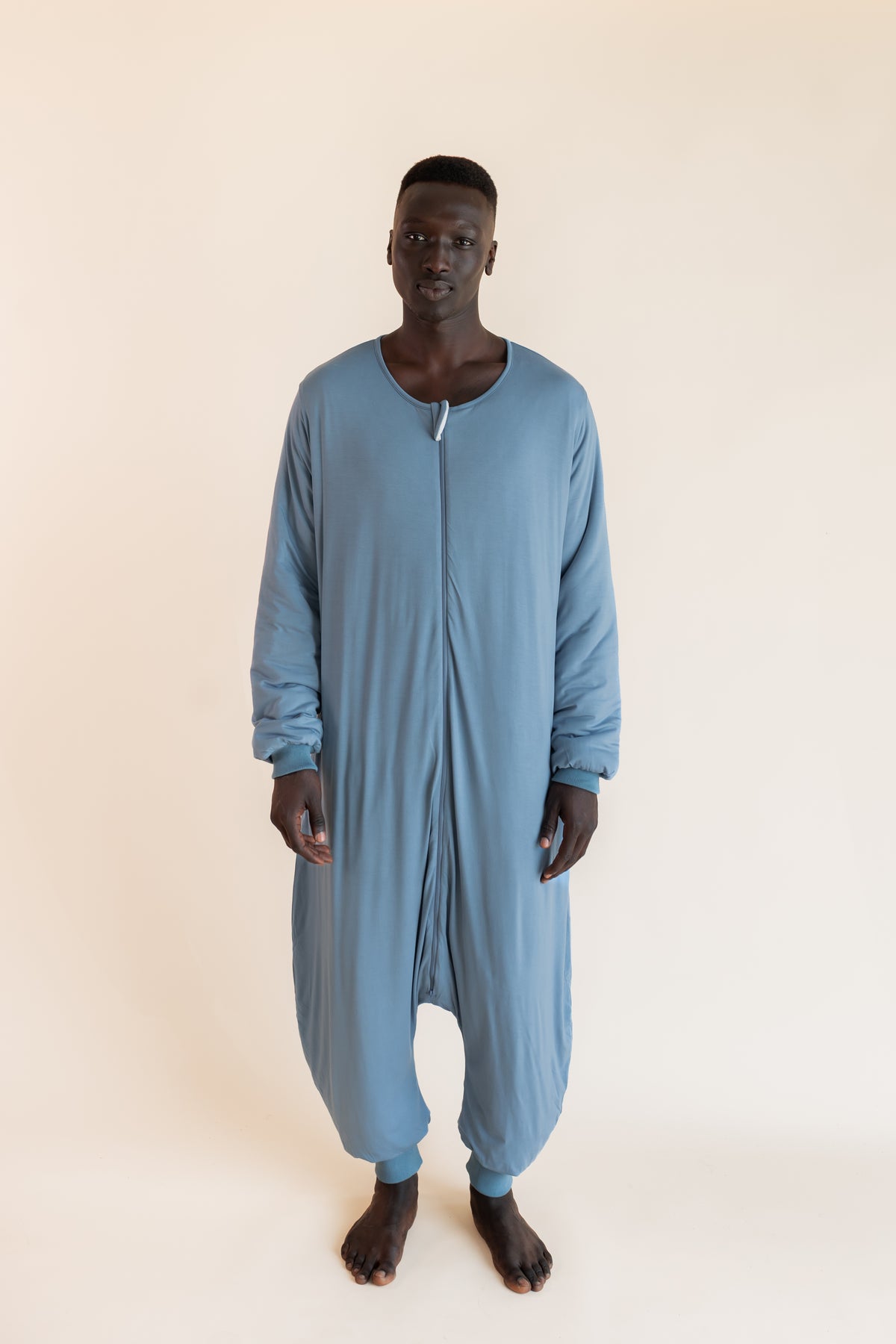 A man wearing a dusty blue, long-sleeve, full-body zip-up harem-style sleep sack against a plain, warm off-white studio background.