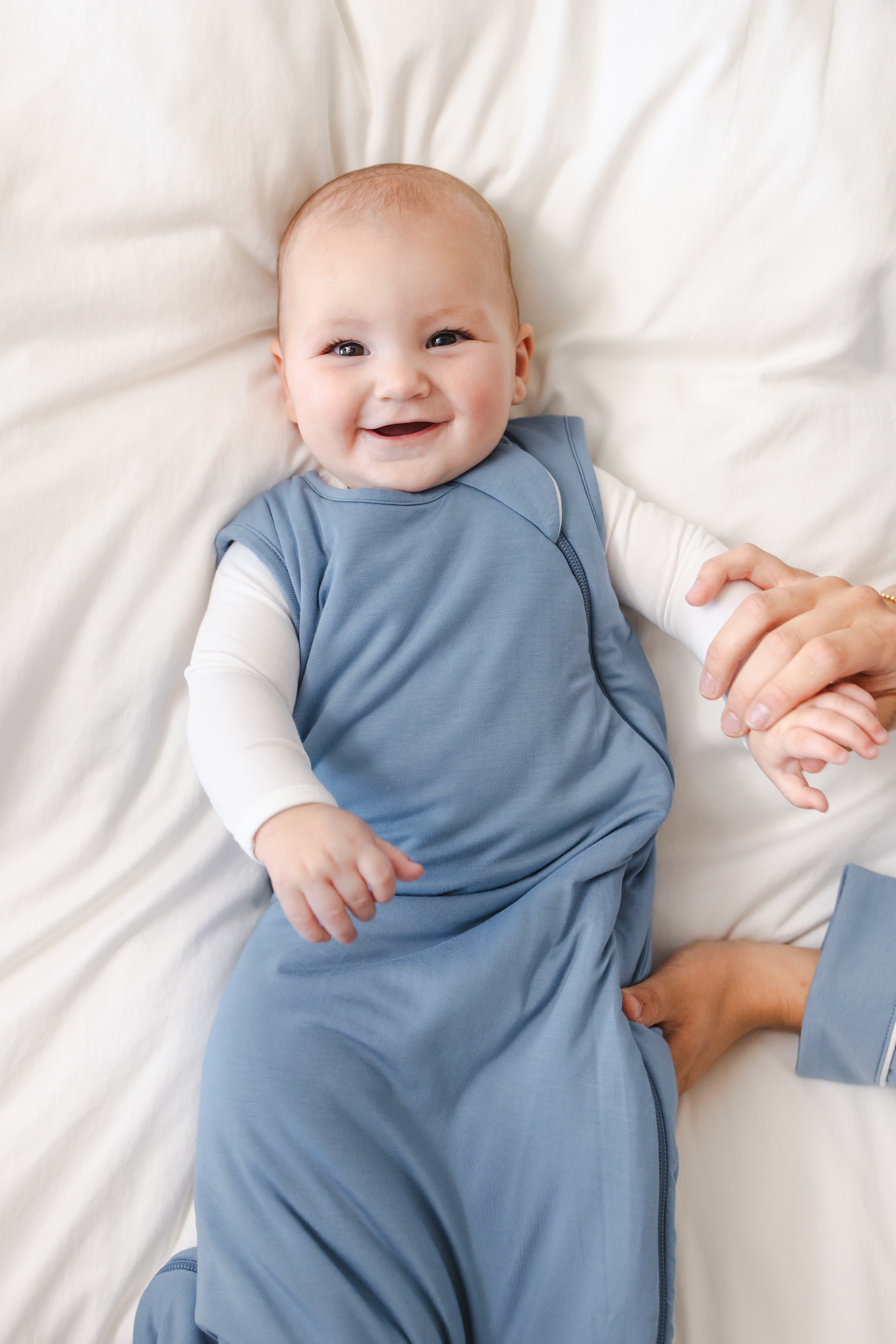 A smiling baby lying on white bedding, wearing a dusty blue sleeveless sleep sack over a white long-sleeve shirt. An adult hand is holding the baby's hand.