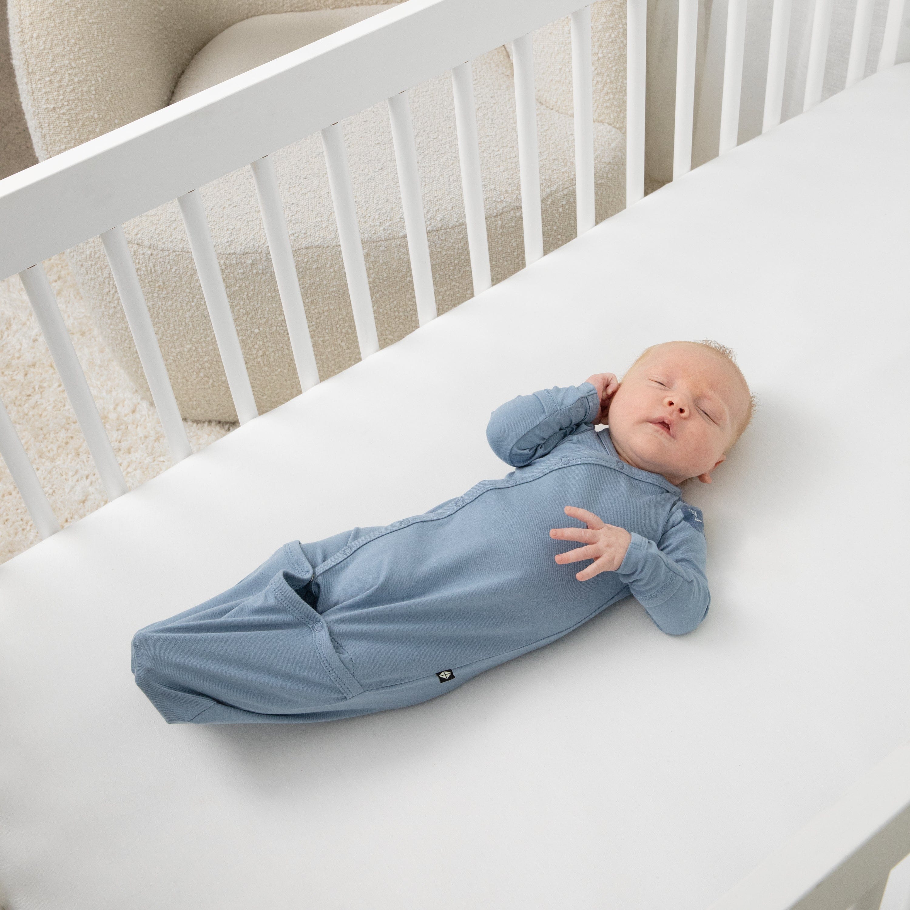Newborn sleeping in a crib on a white crib sheet wearing the Bundler in Slate