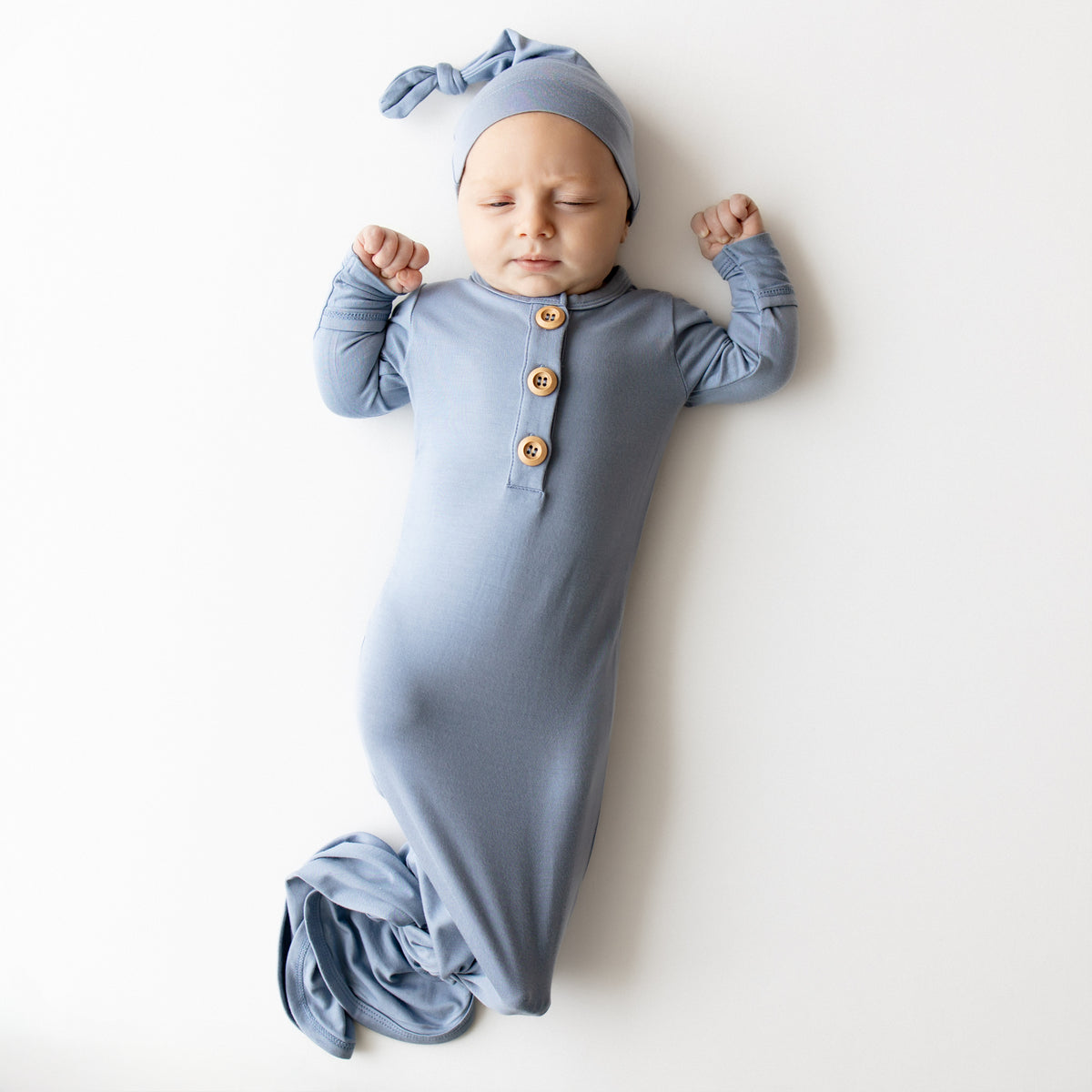 Baby in a knotted gown in a slate blue color that has wooden buttons and matching cap