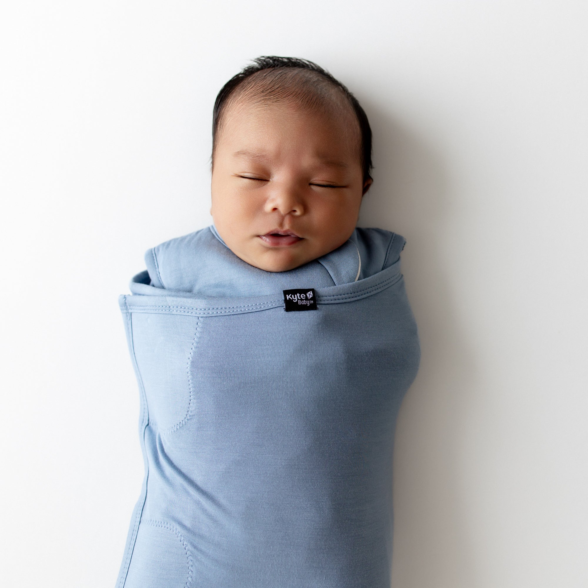 Newborn baby wrapped in a blue swaddler with a Kyte Baby label on a white background
