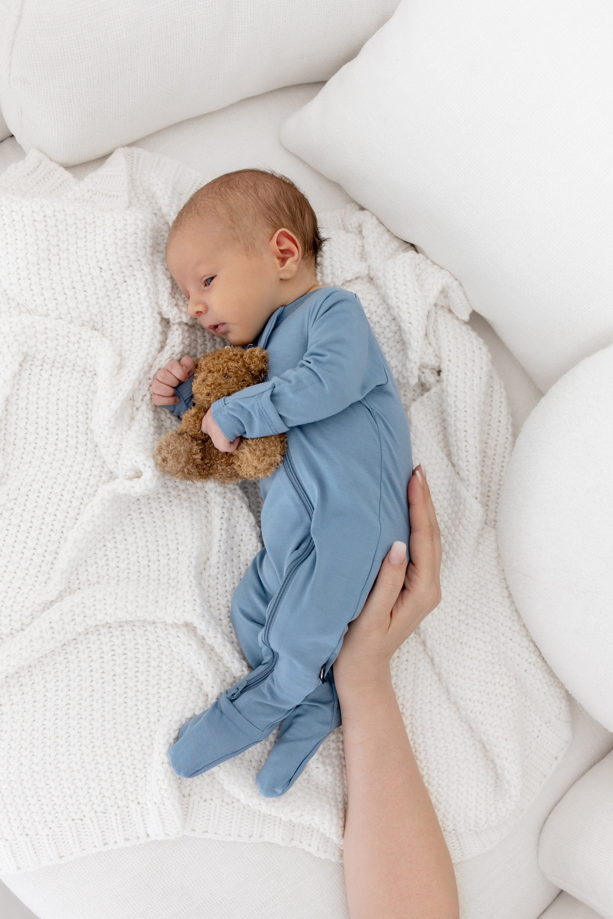 Baby in a blue footie pajama holding a teddy bear, lying on a white blanket.