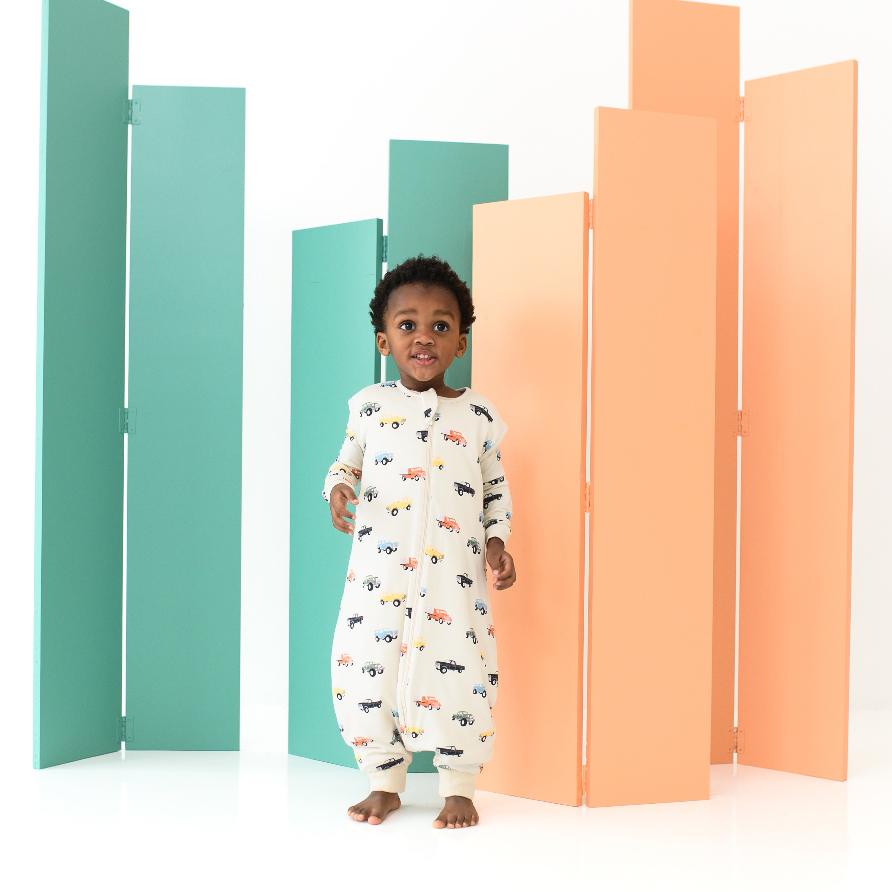 Young model standing wearing the Sleep Bag Walker in Vintage Truck 1.0 with matching long sleeve pajamas underneath
