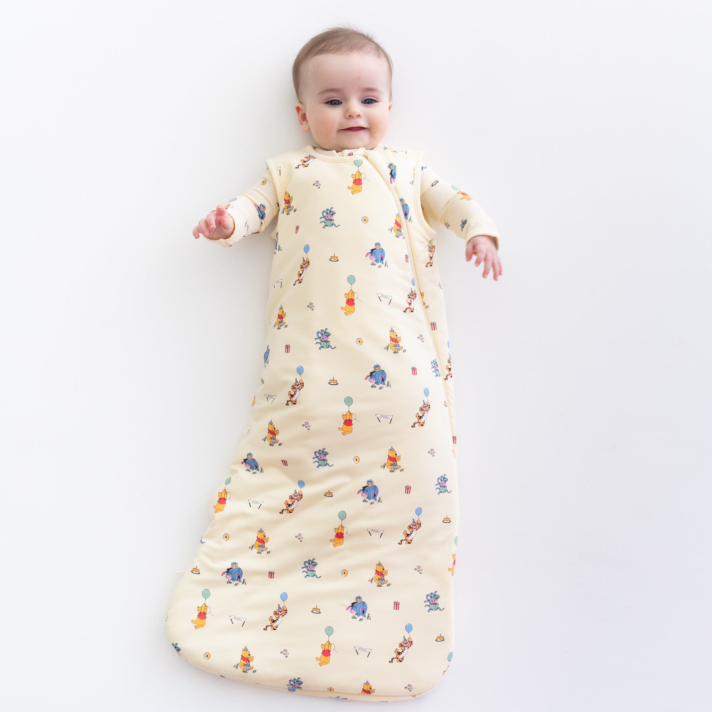 Young infant laying on a white surface wearing the Sleep Bag in Winnie the Pooh Celebration 1.0 with a matching romper on underneath