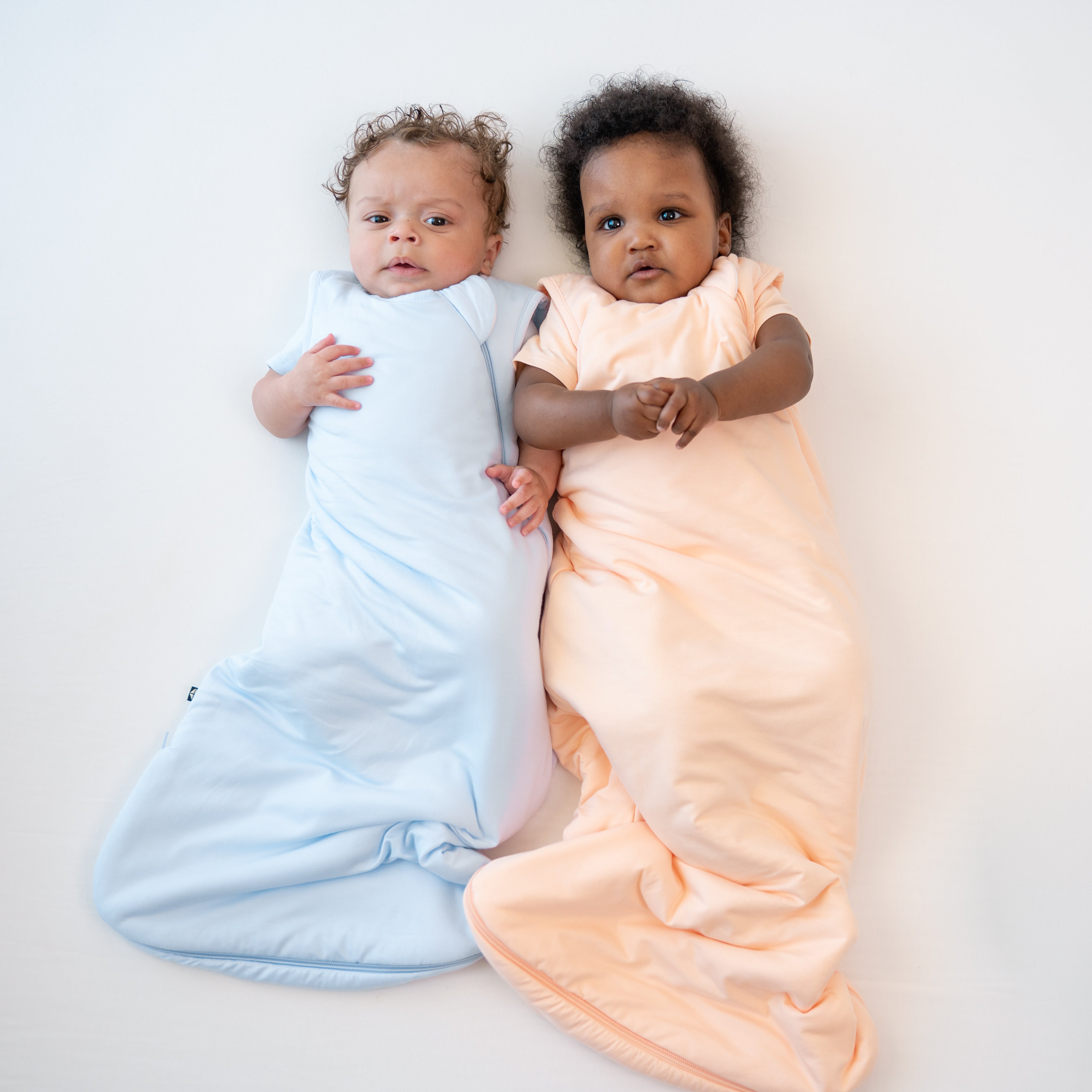 Two infant models laying side by side wearing 1.0 Sleep Bags in Mist and Canyon