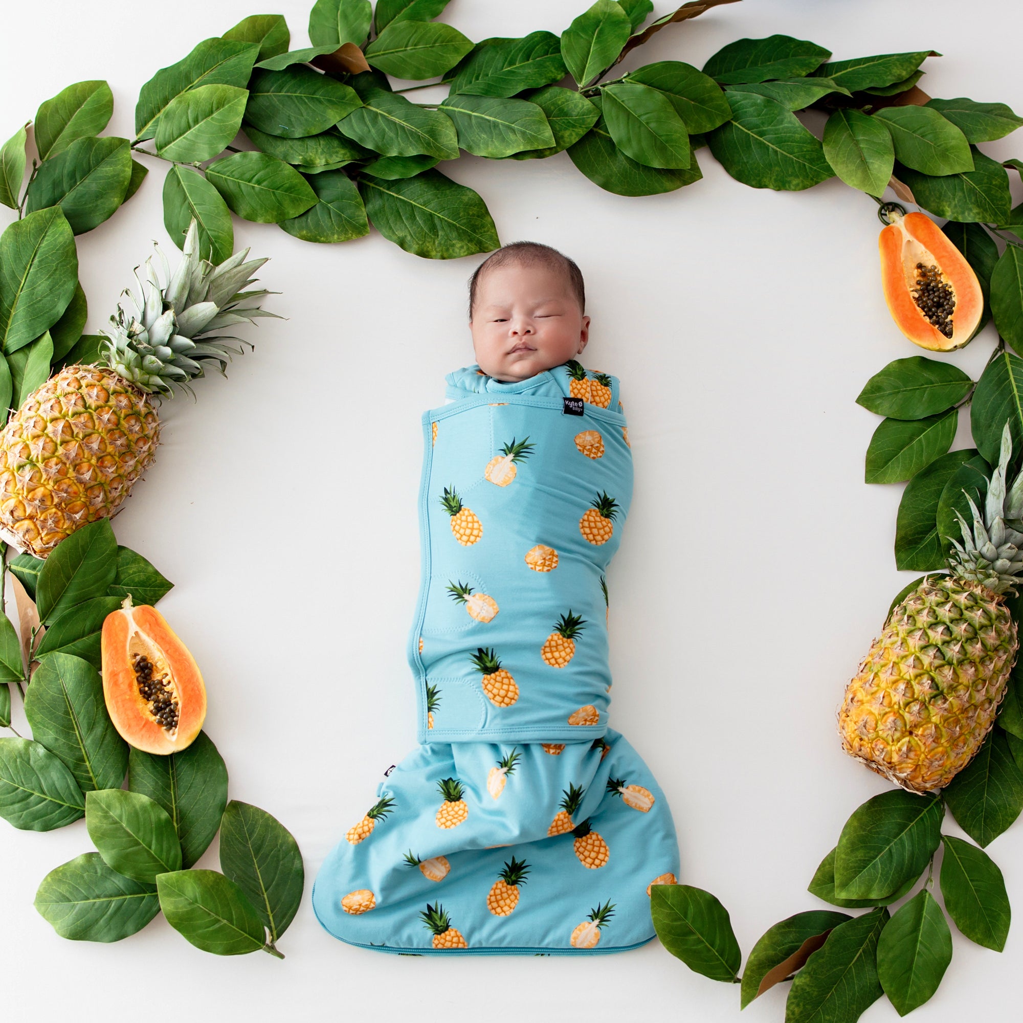 Baby swaddled in Sleep Bag Swaddler in Pineapple 1.0 surrounded by fruit and leaves