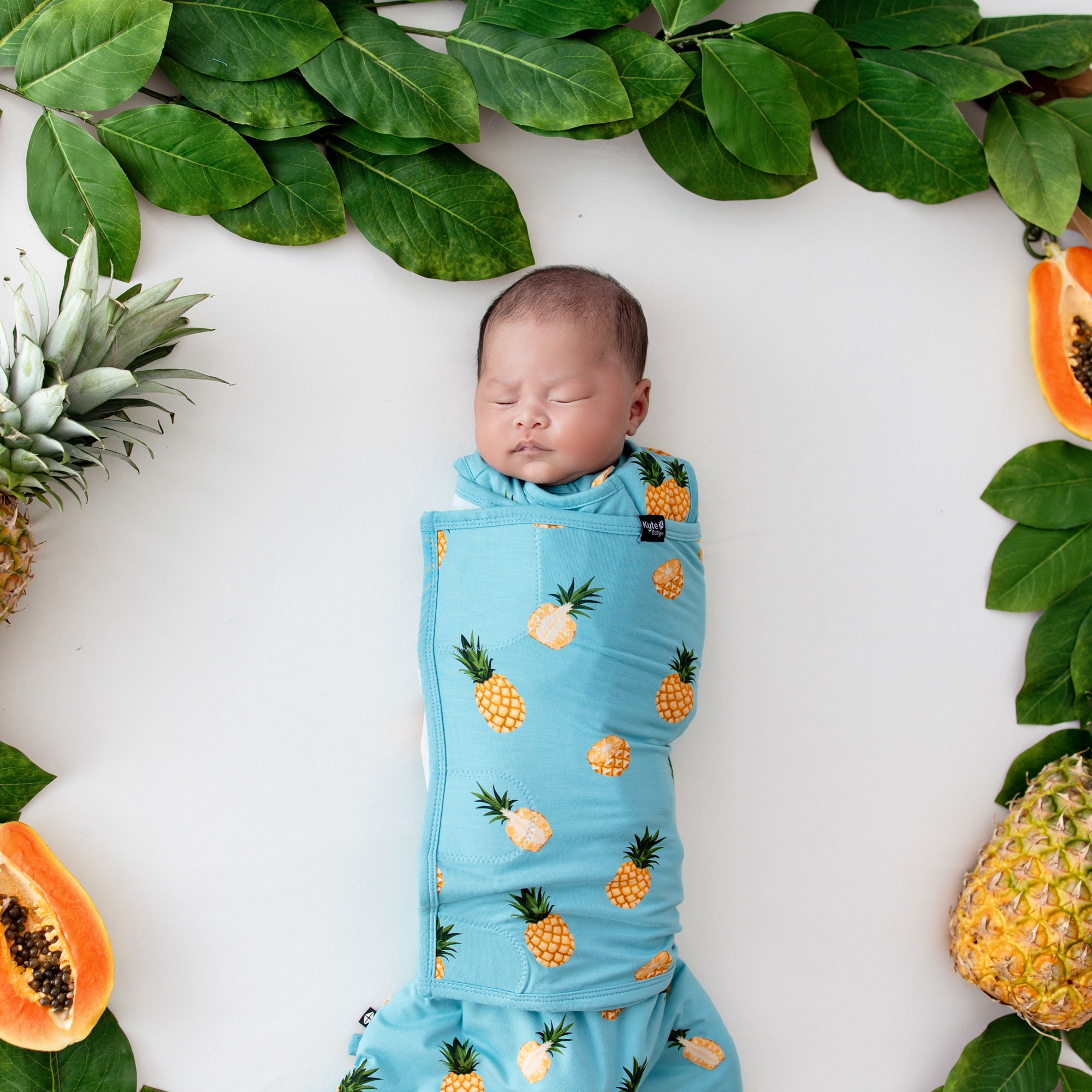 Baby in Sleep Bag Swaddler in Pineapple 1.0 surrounded by fruit and leaves close up