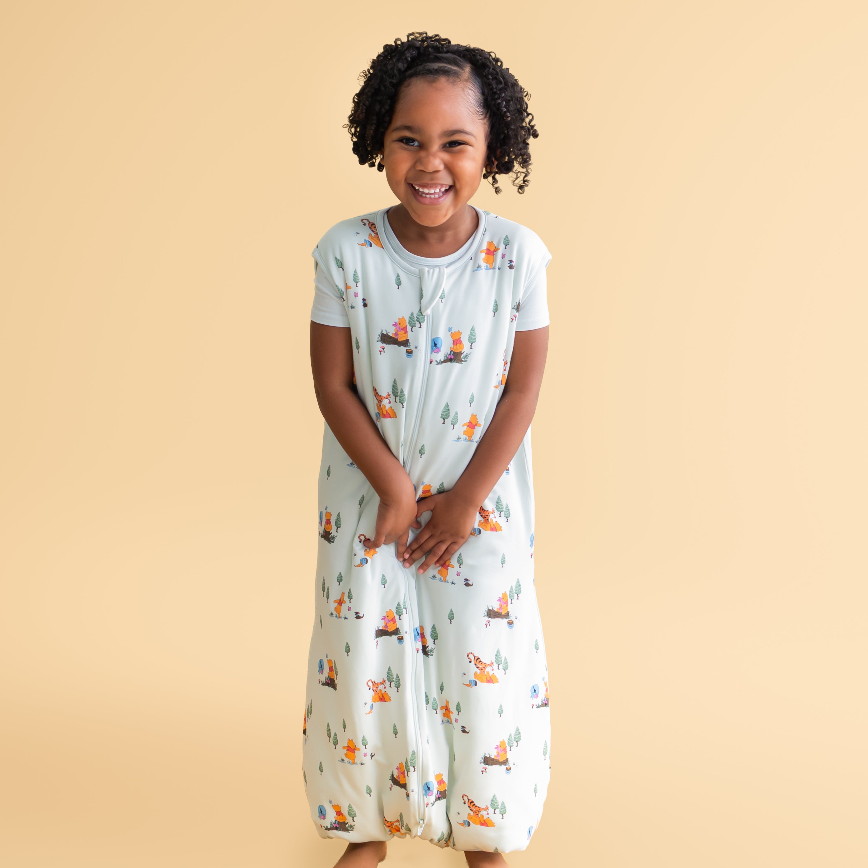 Smiling girl wearing the Sleep Bag Walker in Winnie the Pooh Hundred Acre Wood 1.0 standing in front of a mustard yellow background