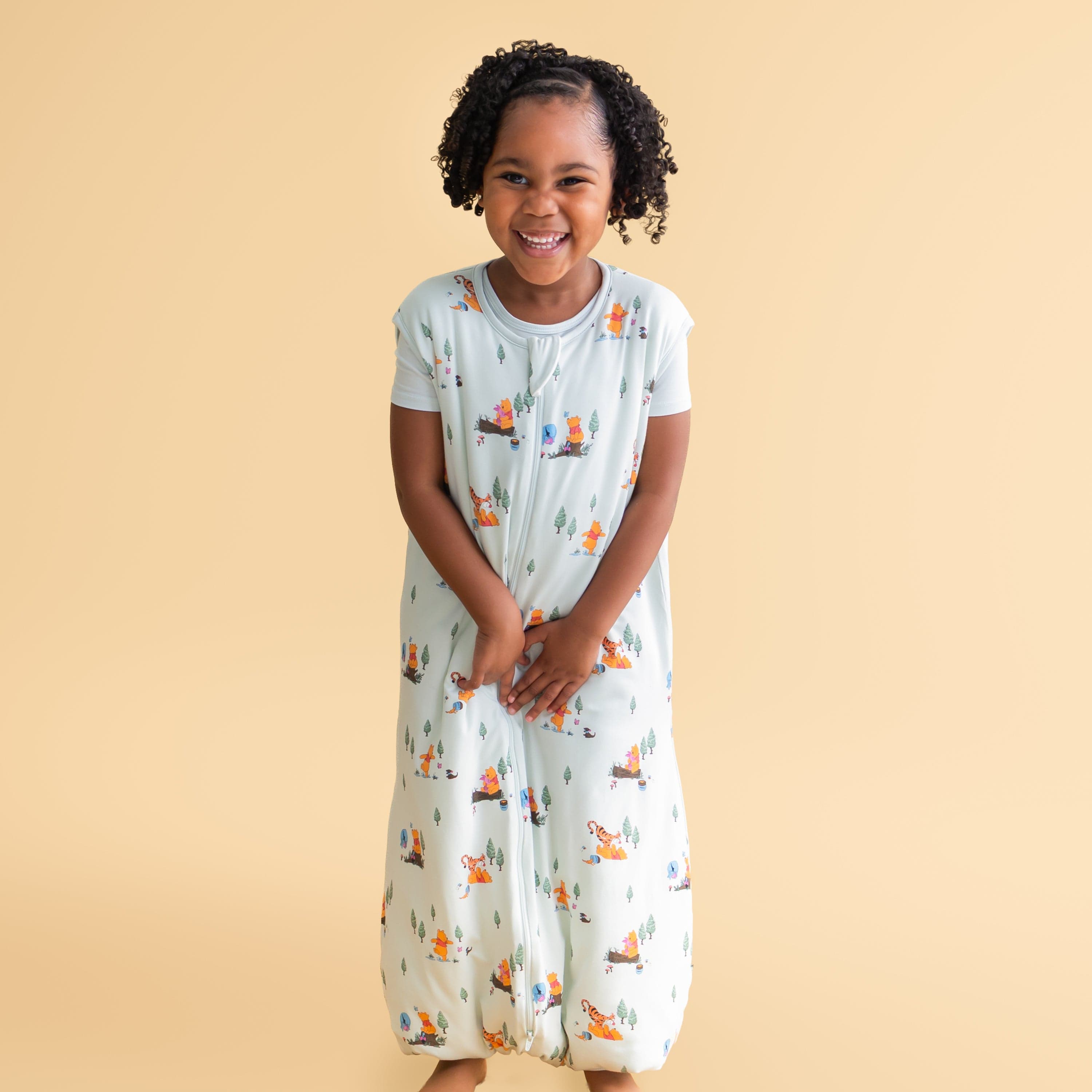 Smiling girl wearing the Sleep Bag Walker in Winnie the Pooh Hundred Acre Wood 1.0 standing in front of a mustard yellow background