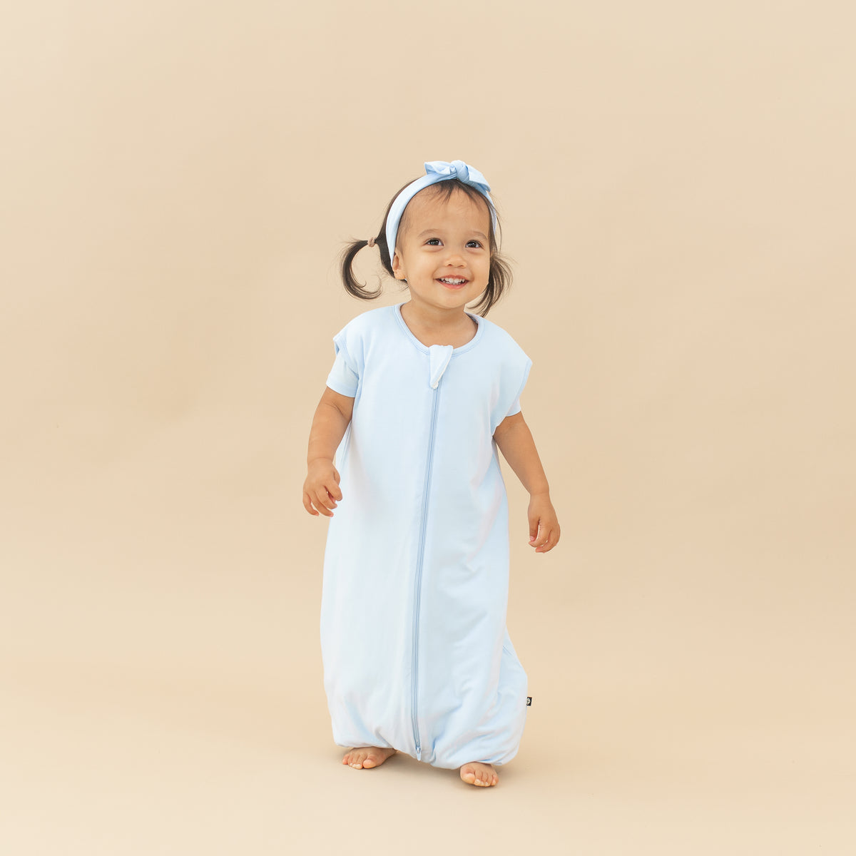 Smiling model walking in the Sleep Bag Walker in Mist 1.0 with matching knotted bow