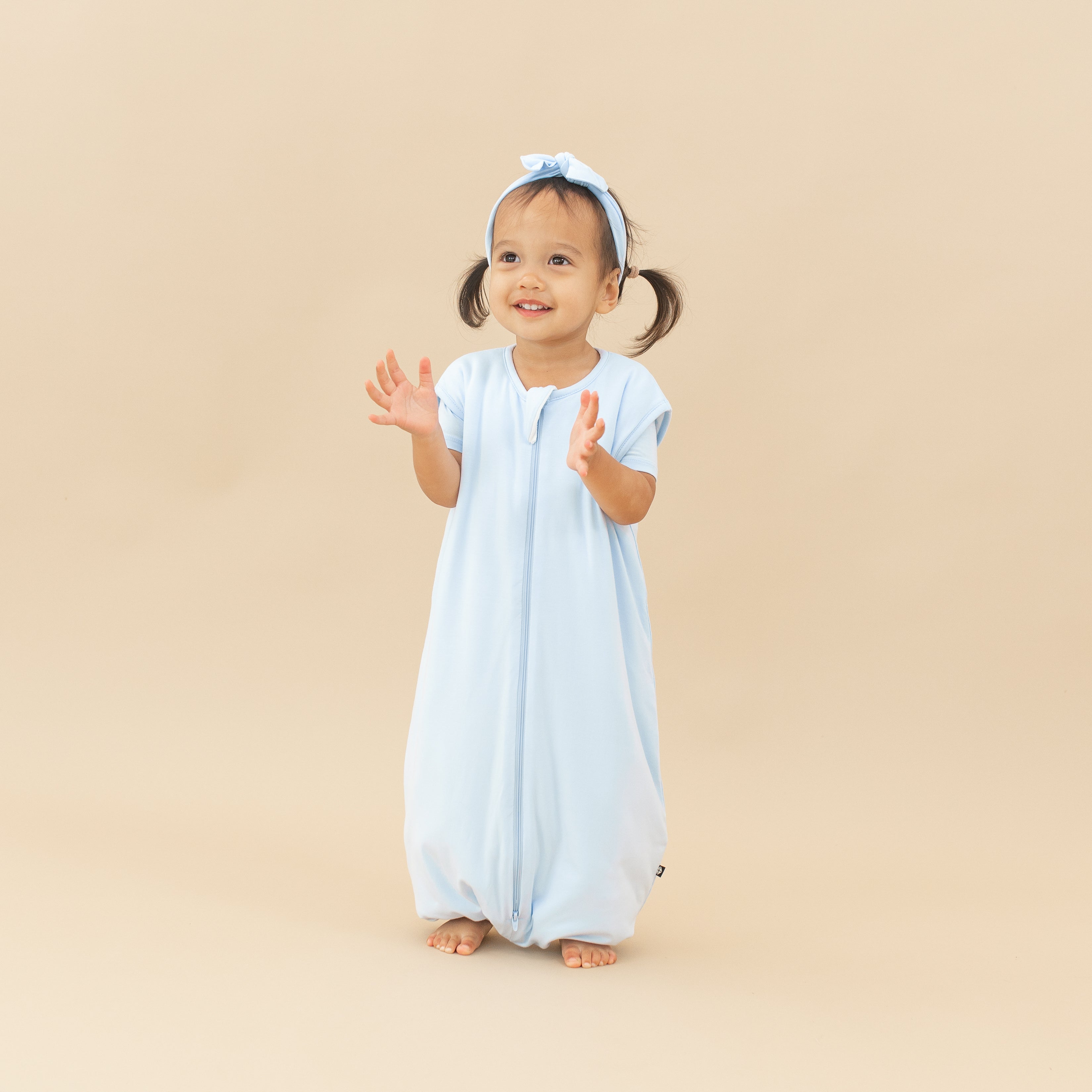 Smiling child model clapping her hands while wearing the Sleep Bag Walker in Mist 1.0 with the matching knotted bow