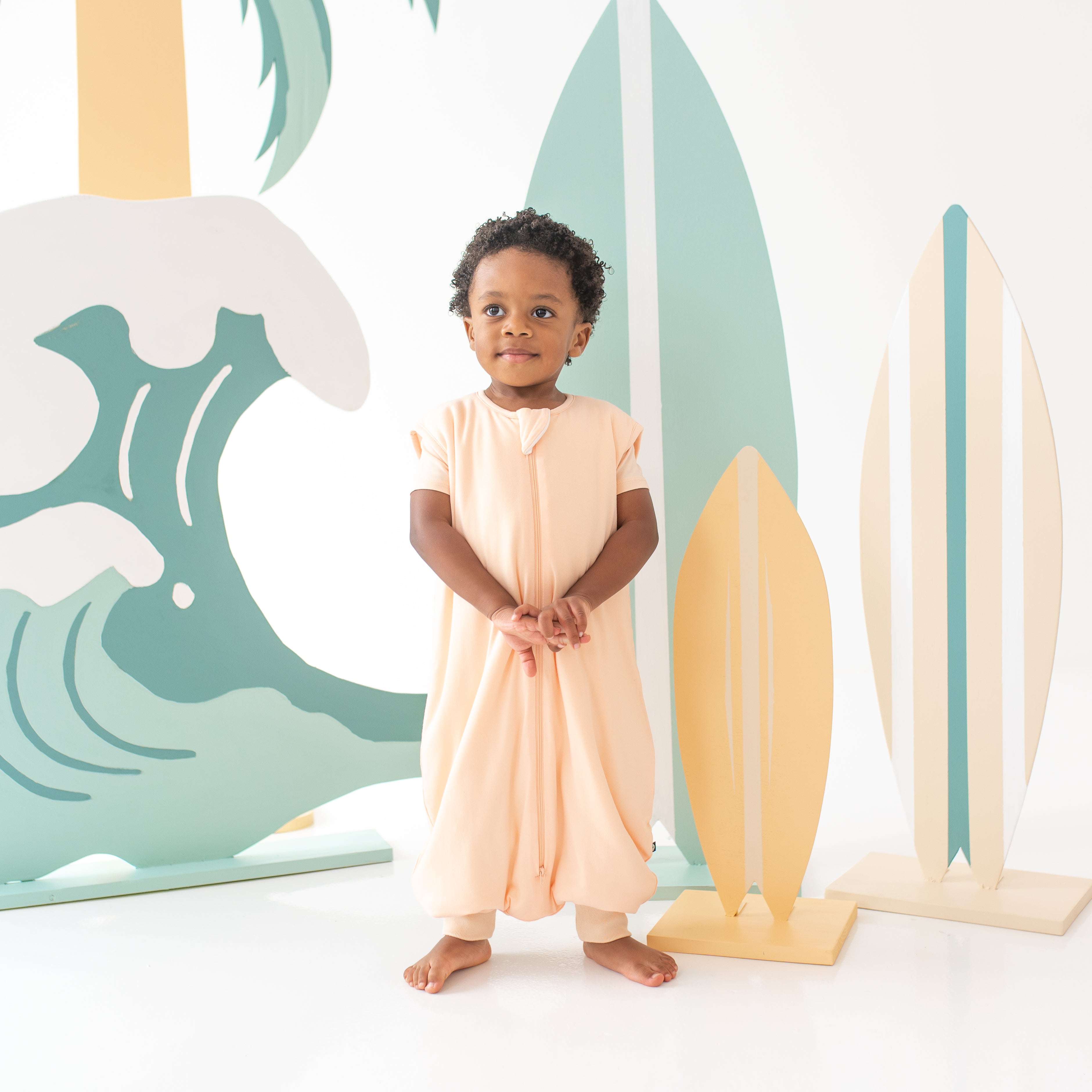 Child model standing clasping hands together wearing the Sleep Bag Walker in Canyon 1.0 with wooden surfboards as a back drop