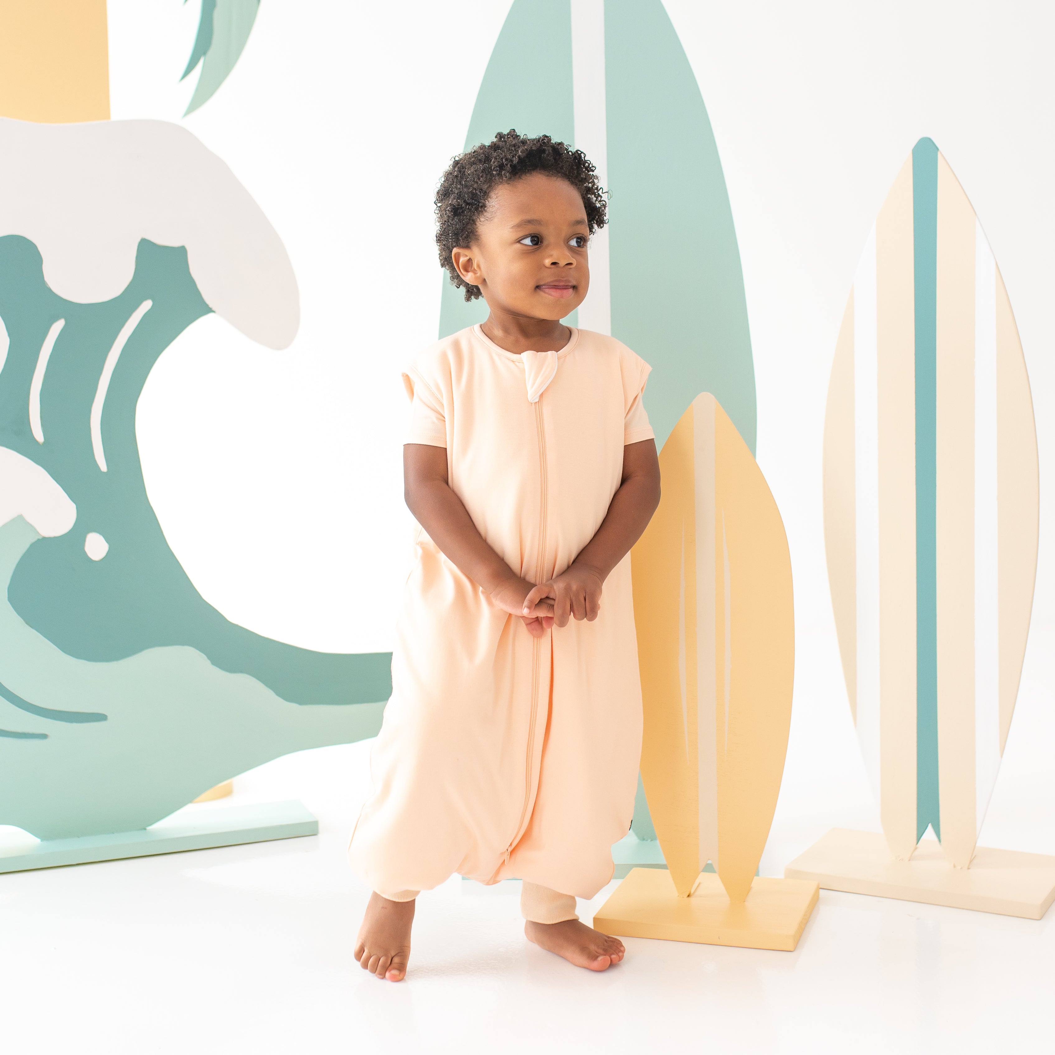 Child model walking clasping hands together wearing the Sleep Bag Walker in Canyon 1.0 with a surfboard backdrop
