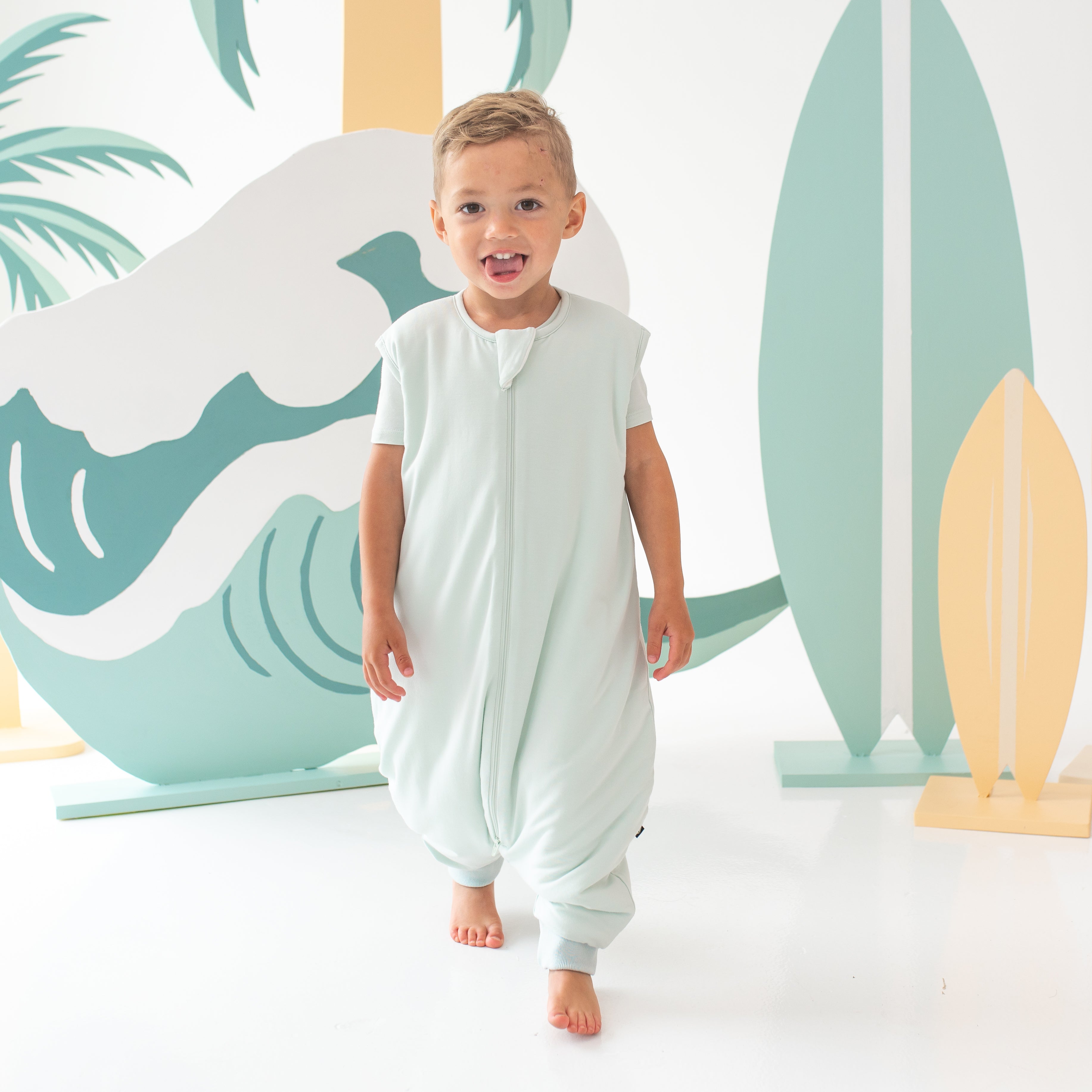 Young child model walking and sticking his tongue out wearing the Sleep Bag Walker in Dew 1.0