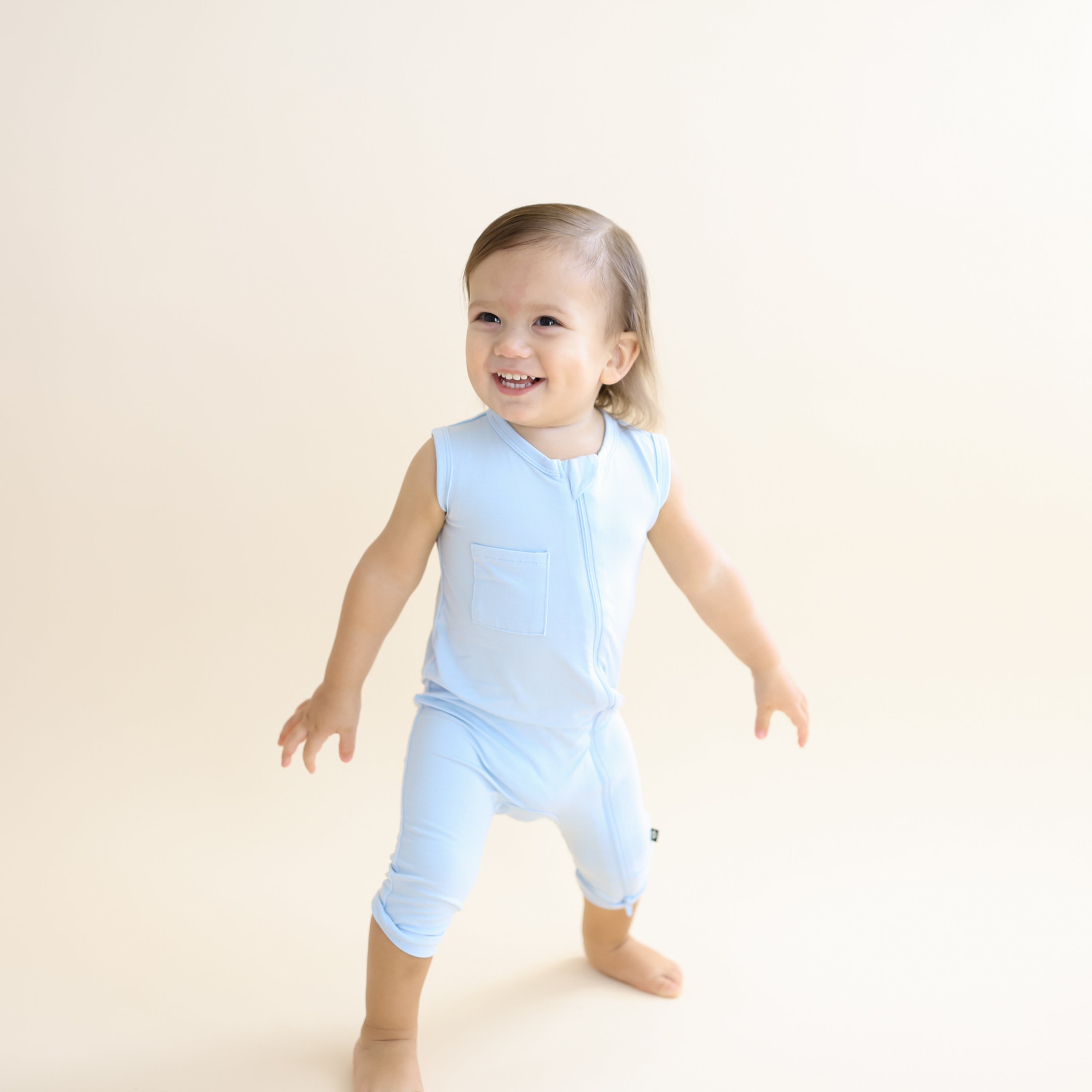 Young smiling boy standing wearing the Zippered Sleeveless Romper in Breeze in front of a light neutral background