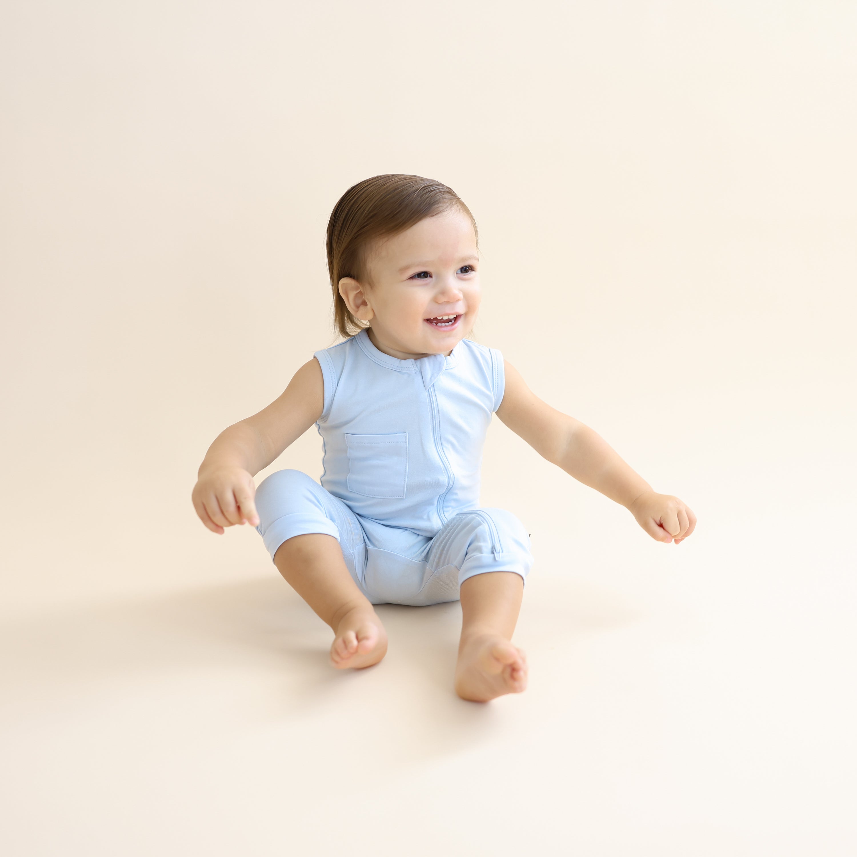 Young toddler sitting on the floor wearing the Zippered Sleeveless Romper in Breeze