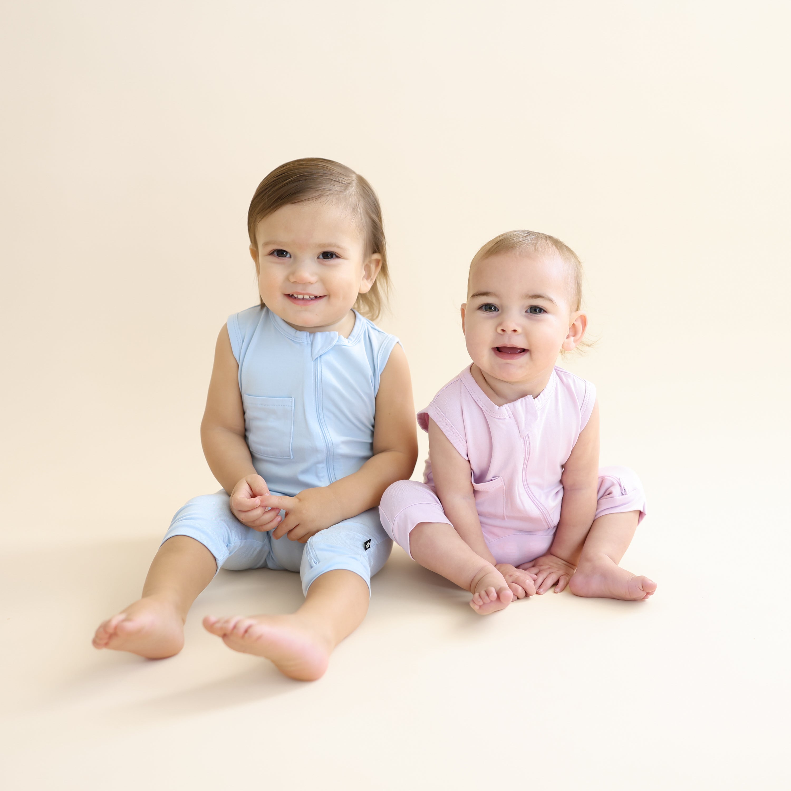 Two young toddlers sitting side by side wearing the Zippered Sleeveless Romper in Breeze and Thistle