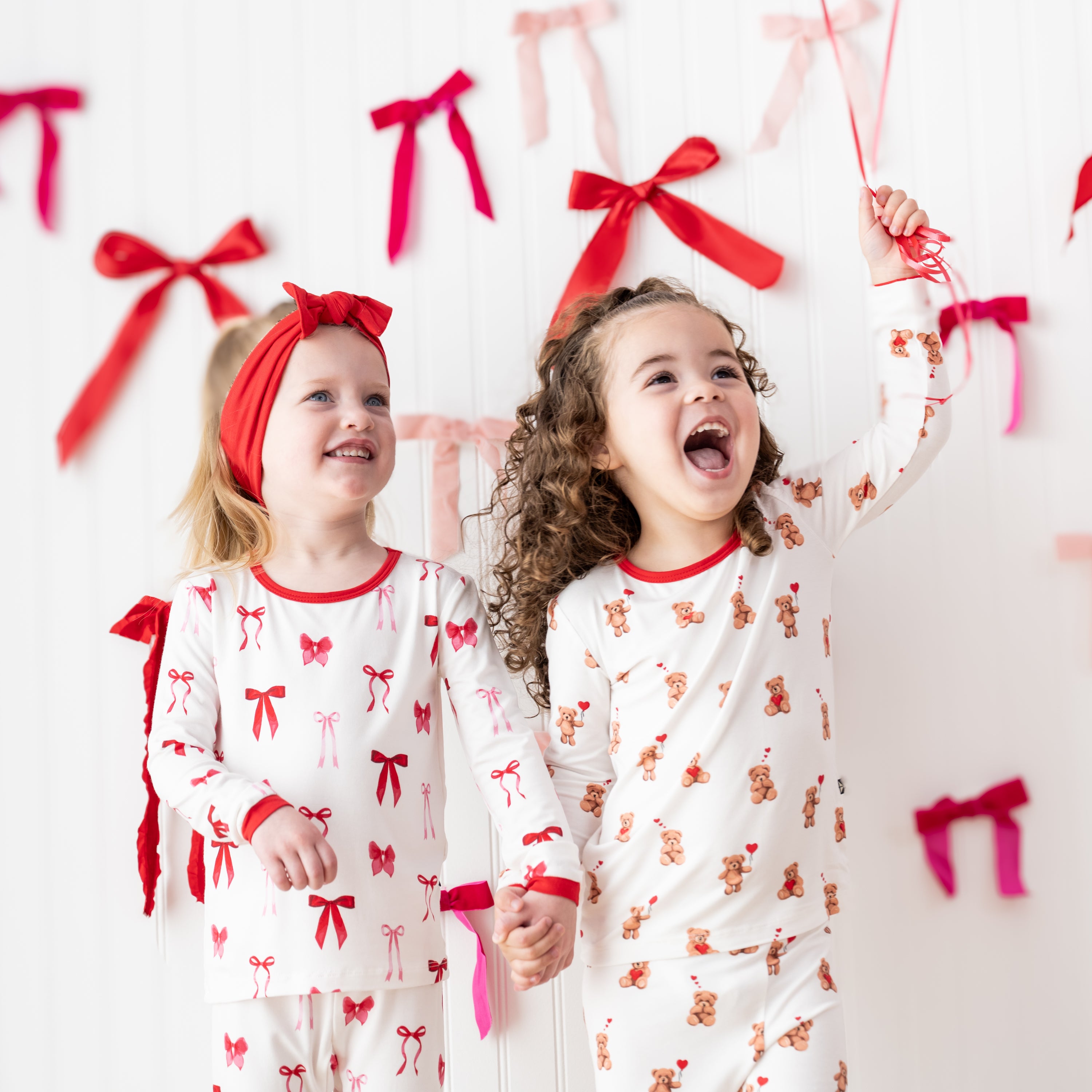 Two girls standing in front of a white paneled wall with bows on it wearing the Long Sleeve Pajamas in Small Love Bow and Bear Hearts