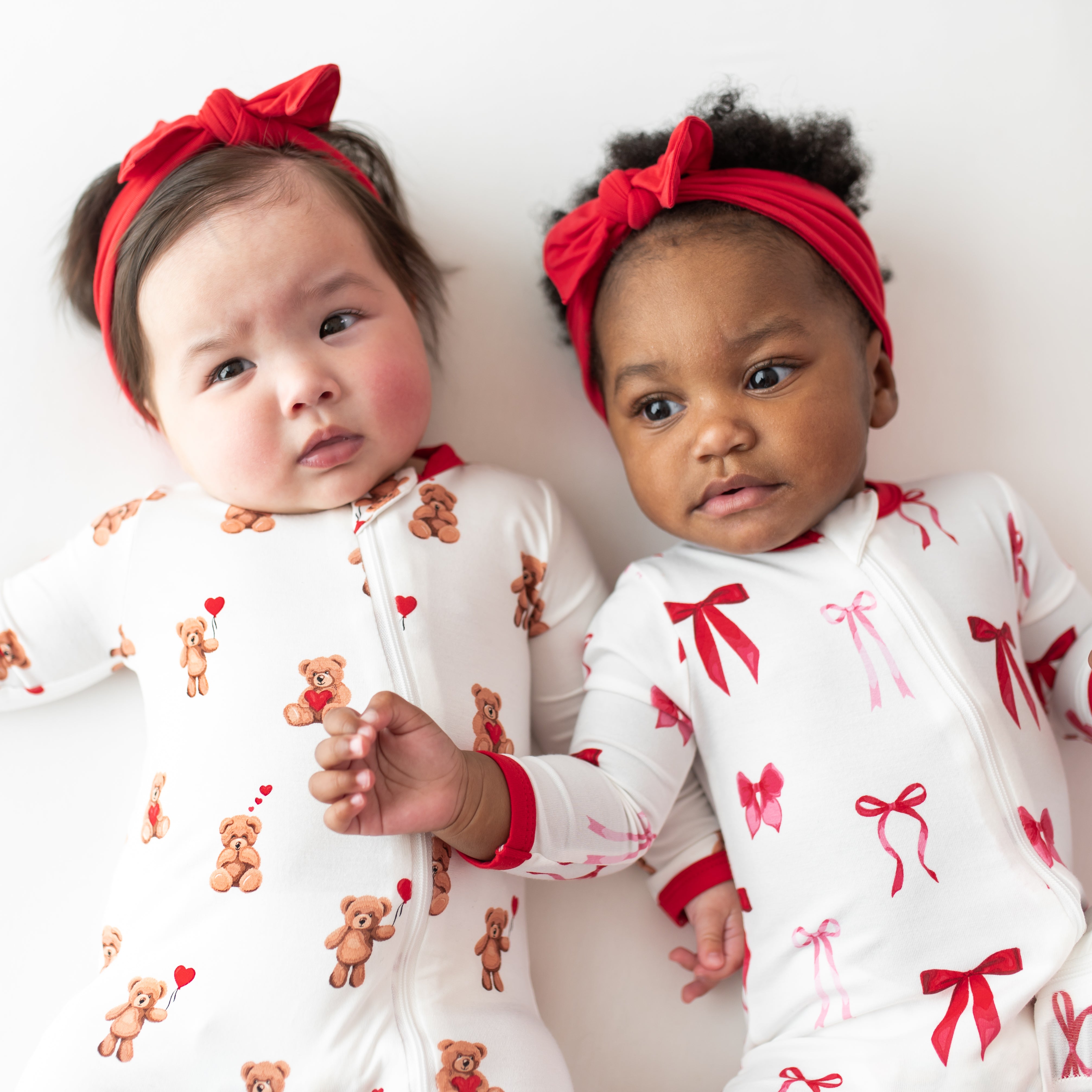 Two toddlers laying side by side wearing the Zippered Romper in Bear Hearts and Small love bow with knotted bow headbands in cardinal