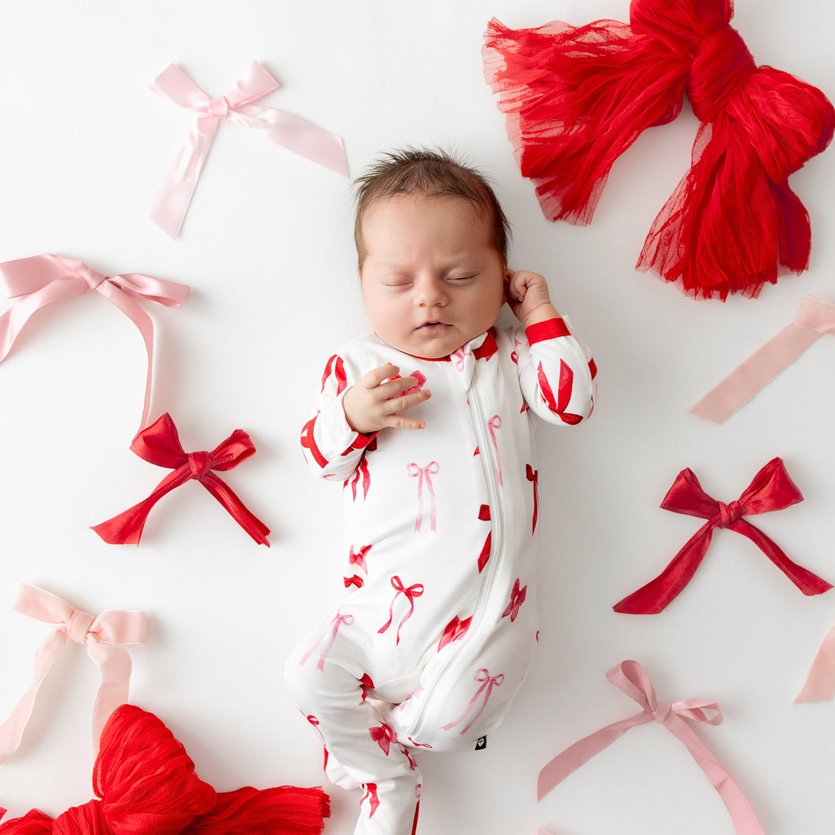Sleeping infant wearing the Zippered Footie in Small Love Bow surrounded by various bows