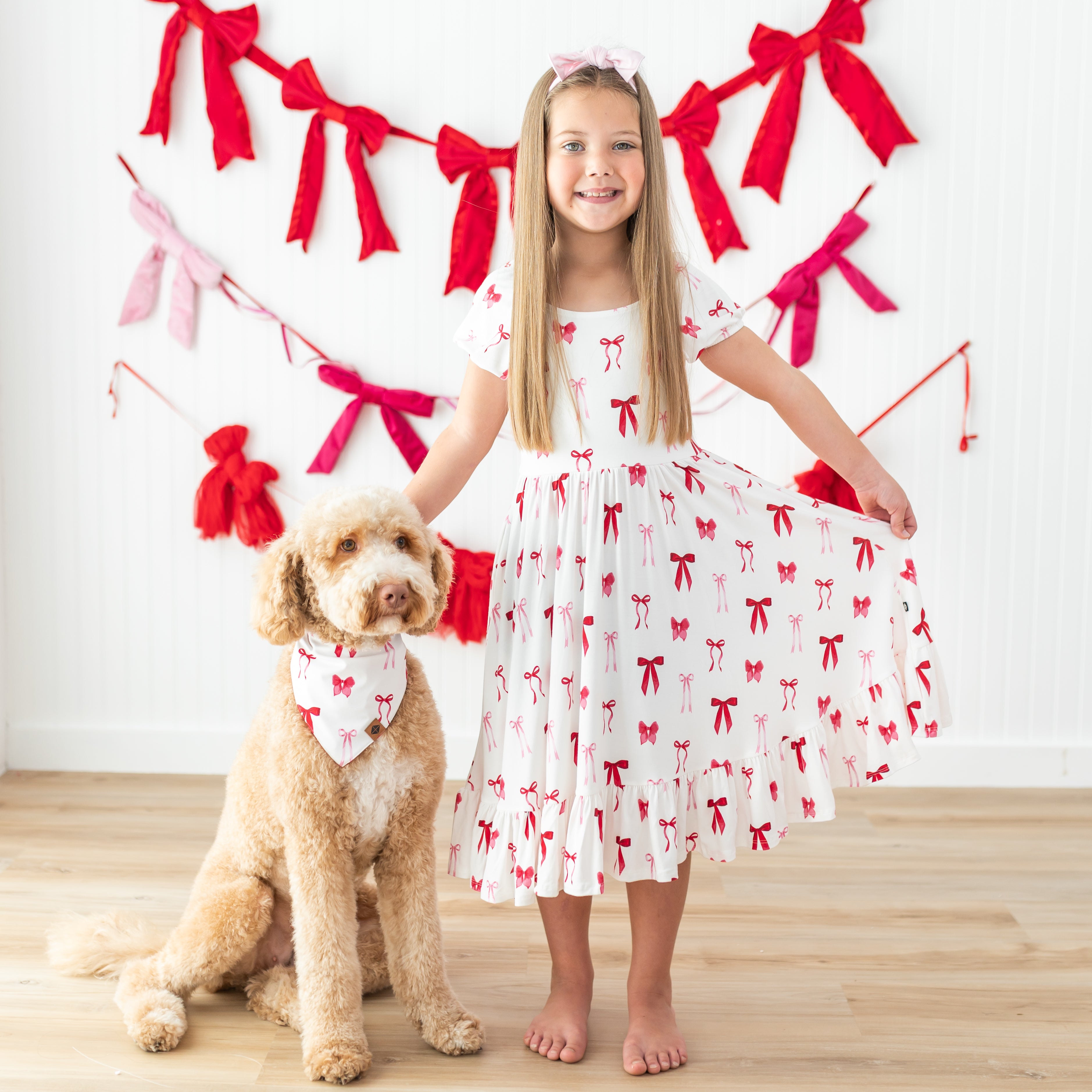 Smiling girl wearing the Puff Sleeve Twirl Dress in Small Love Bow standing beside her dog who is wearing the matching dog bandana