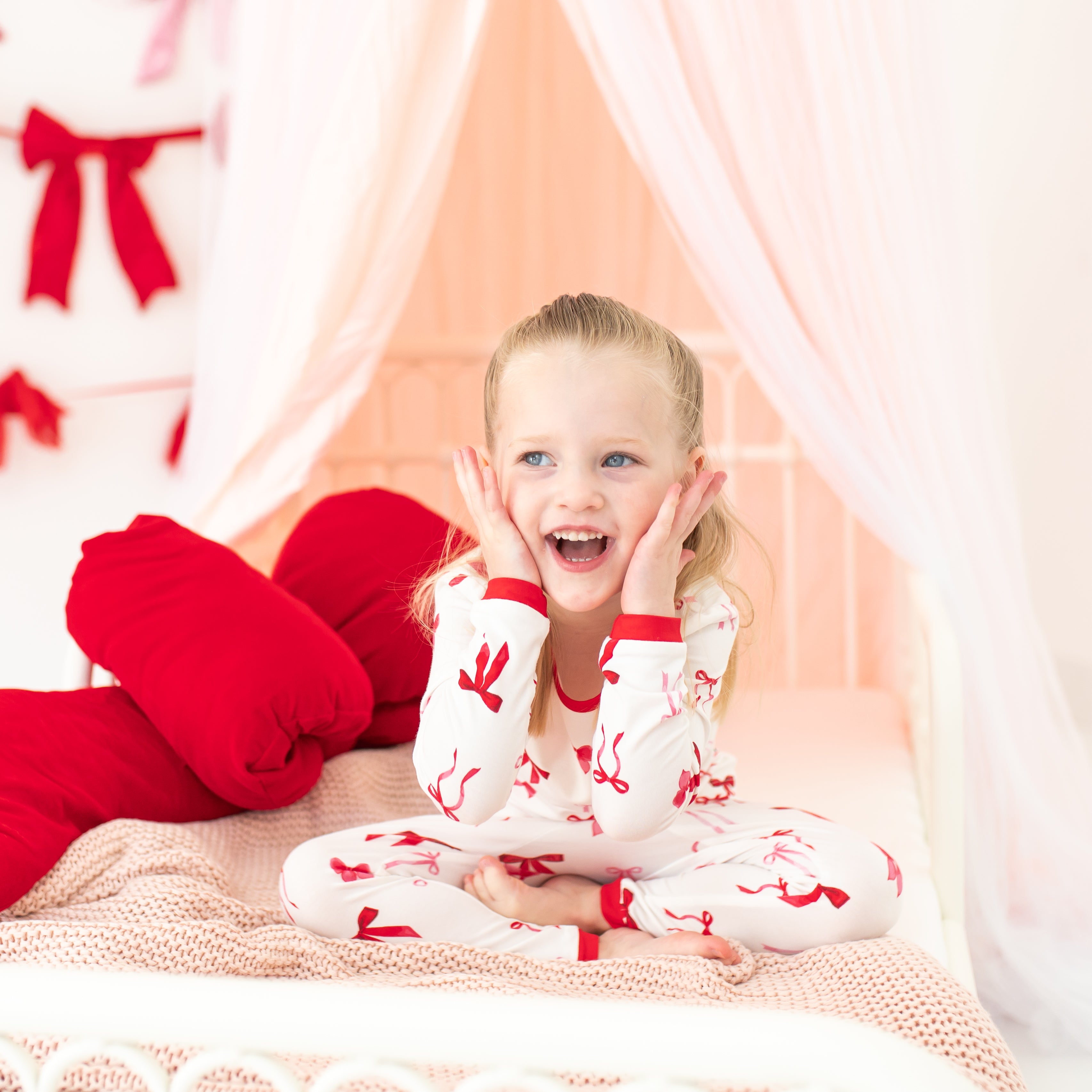 Young girl sitting on a bed wearing the Long Sleeve Pajamas in Small Love Bow with both her hands on her face