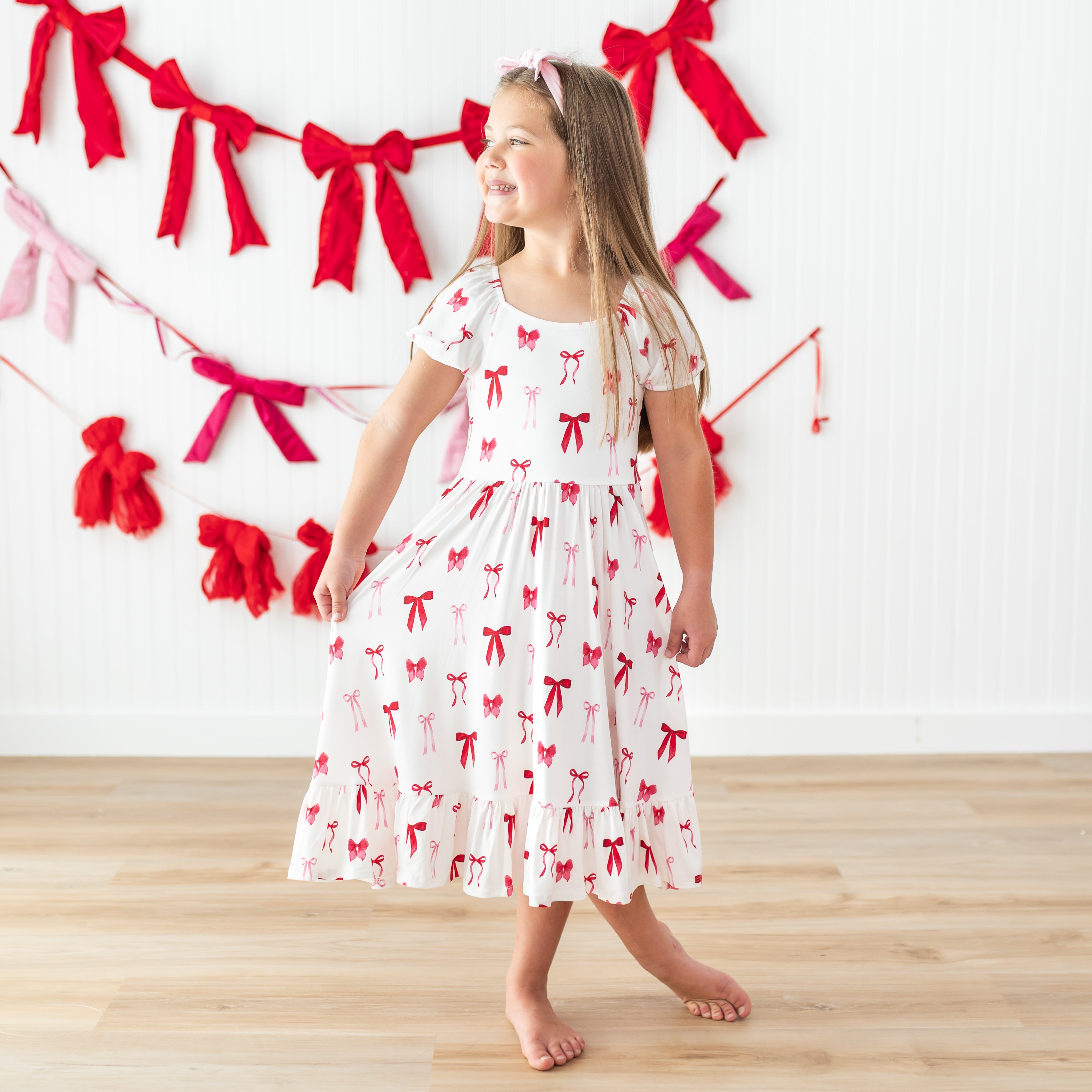 Young girl posing wearing the Puff Sleeve Twirl Dress in Small Love Bow standing in front of a white paneled wall