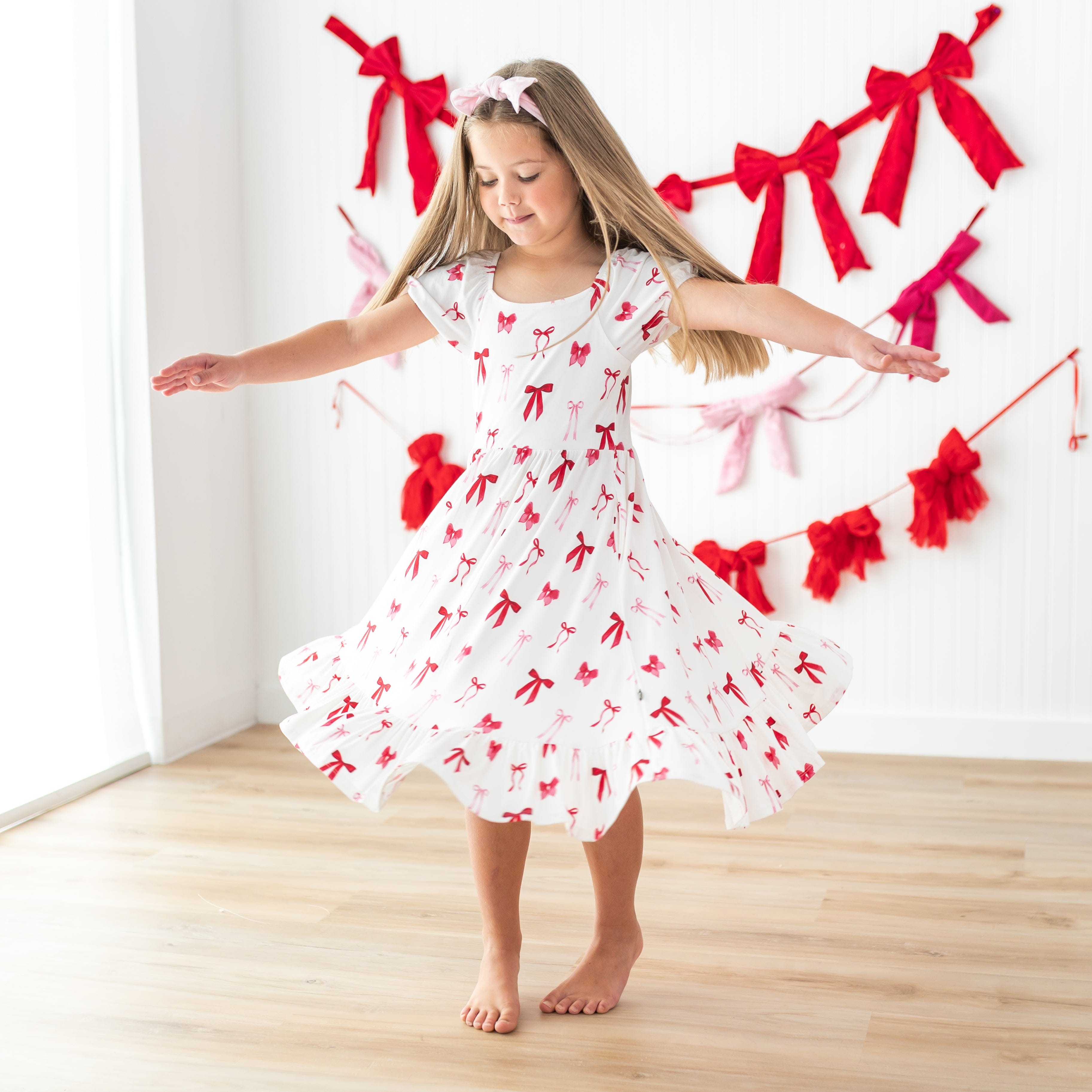 Young girl twirling wearing the flowy Puff Sleeve Twirl Dress in Small Love Bow in front of a white paneled wall with hanging bow garlands