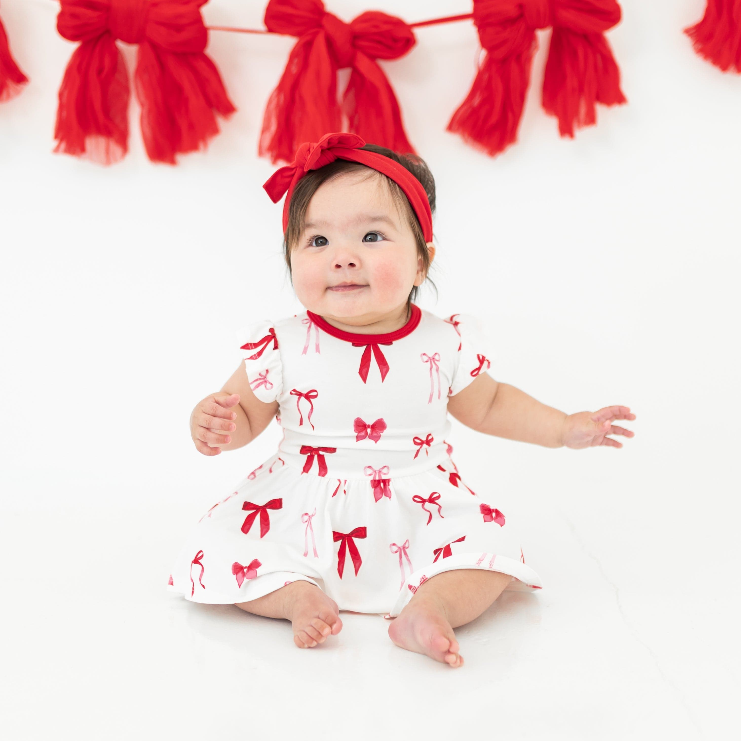 Infant sitting on a cream surface wearing the Twirl Bodysuit Dress in Small Love Bow paired with a Cardinal knotted bow headband