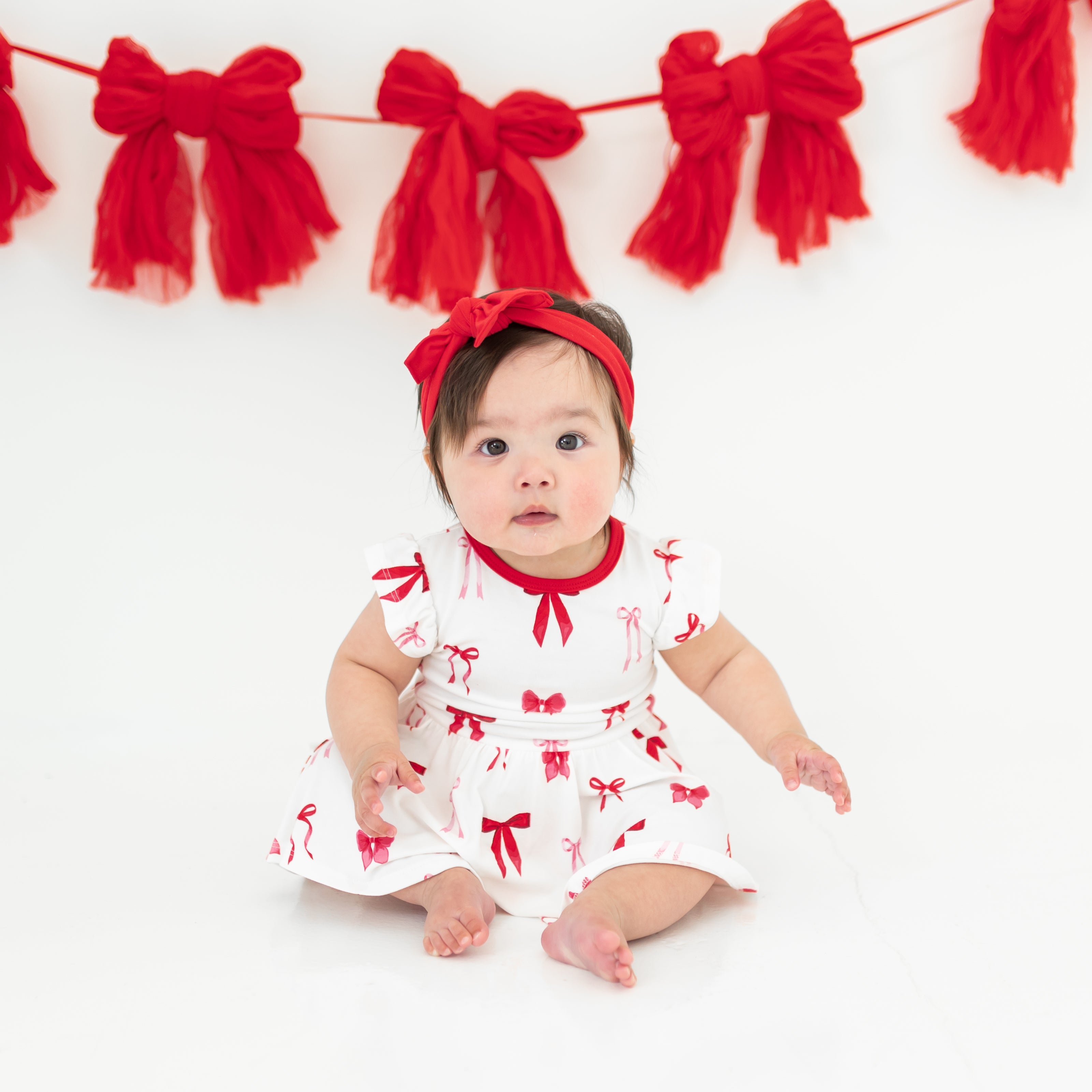 Infant sitting on a light neutral surface wearing the soft and breathable Twirl Bodysuit Dress in Small Love Bow paired with a red bow