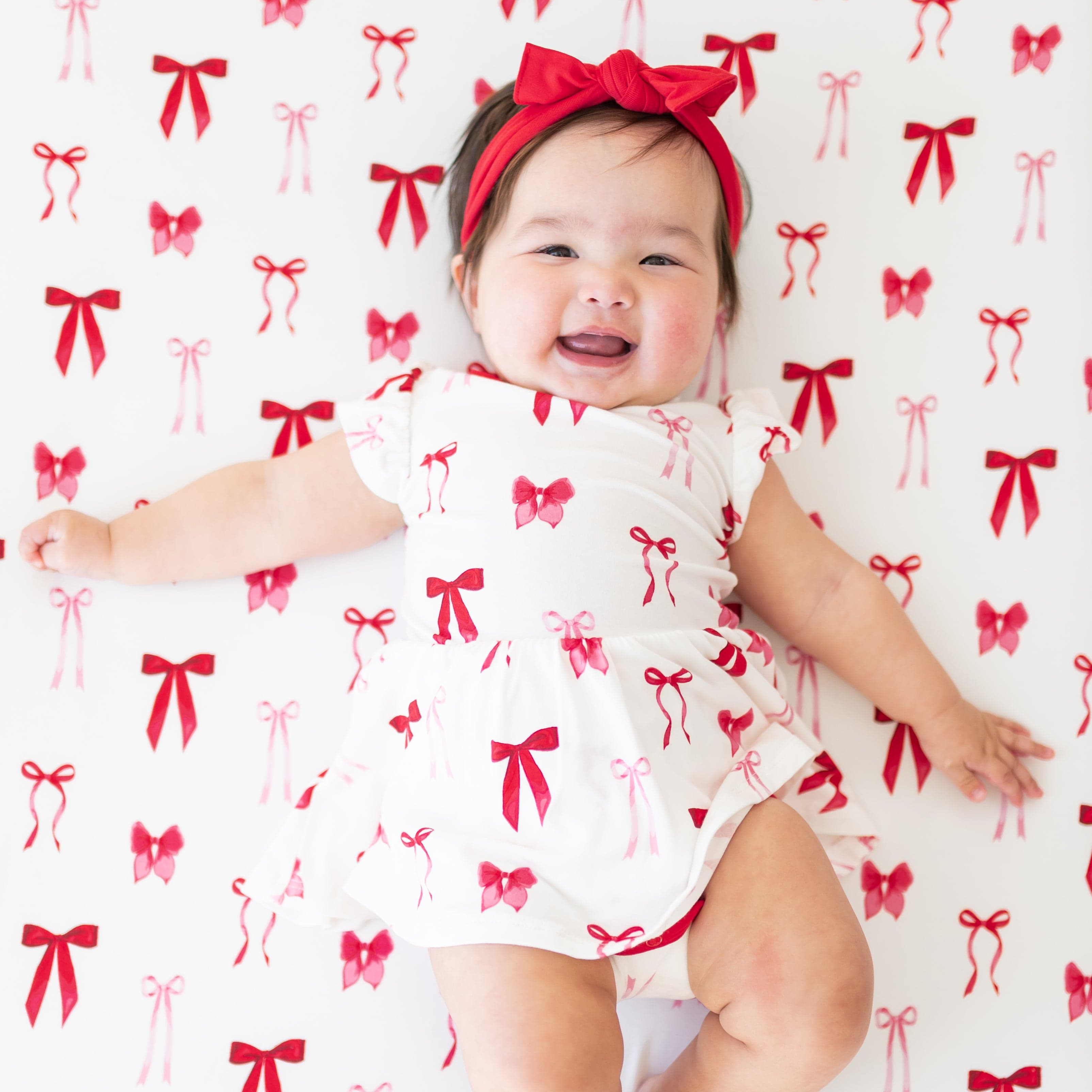 Smiling infant laying in a crib on a Small Love Bow Crib Sheet wearing the Twirl Bodysuit Dress in Small Love Bow paired with a knotted bow headband in cardinal
