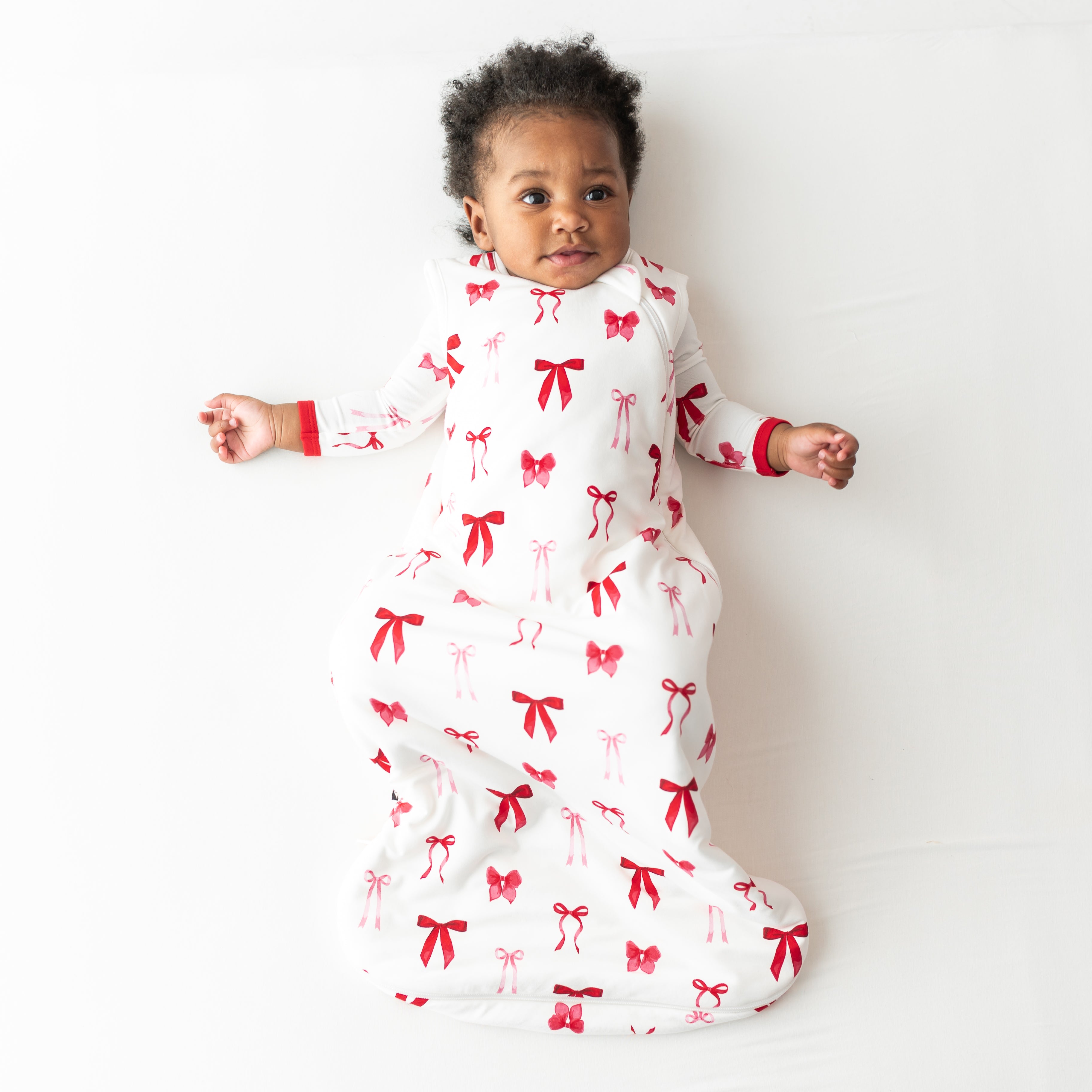 young toddler laying on a light neutral surface wearing the soft and breathable Sleep Bag in Small Love Bow 1.0 with a matching romper on underneath