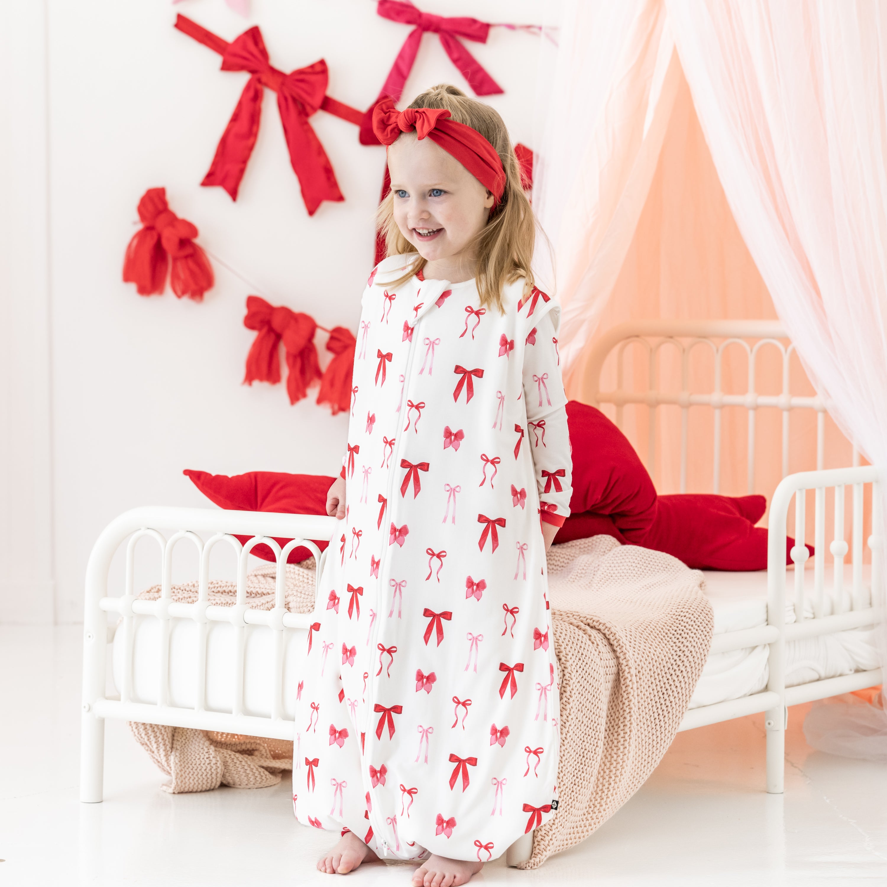 Young girl standing at the end of a bed wearing the Sleep Bag Walker in Small Love Bow 1.0 with matching long sleeve pajamas on underneath