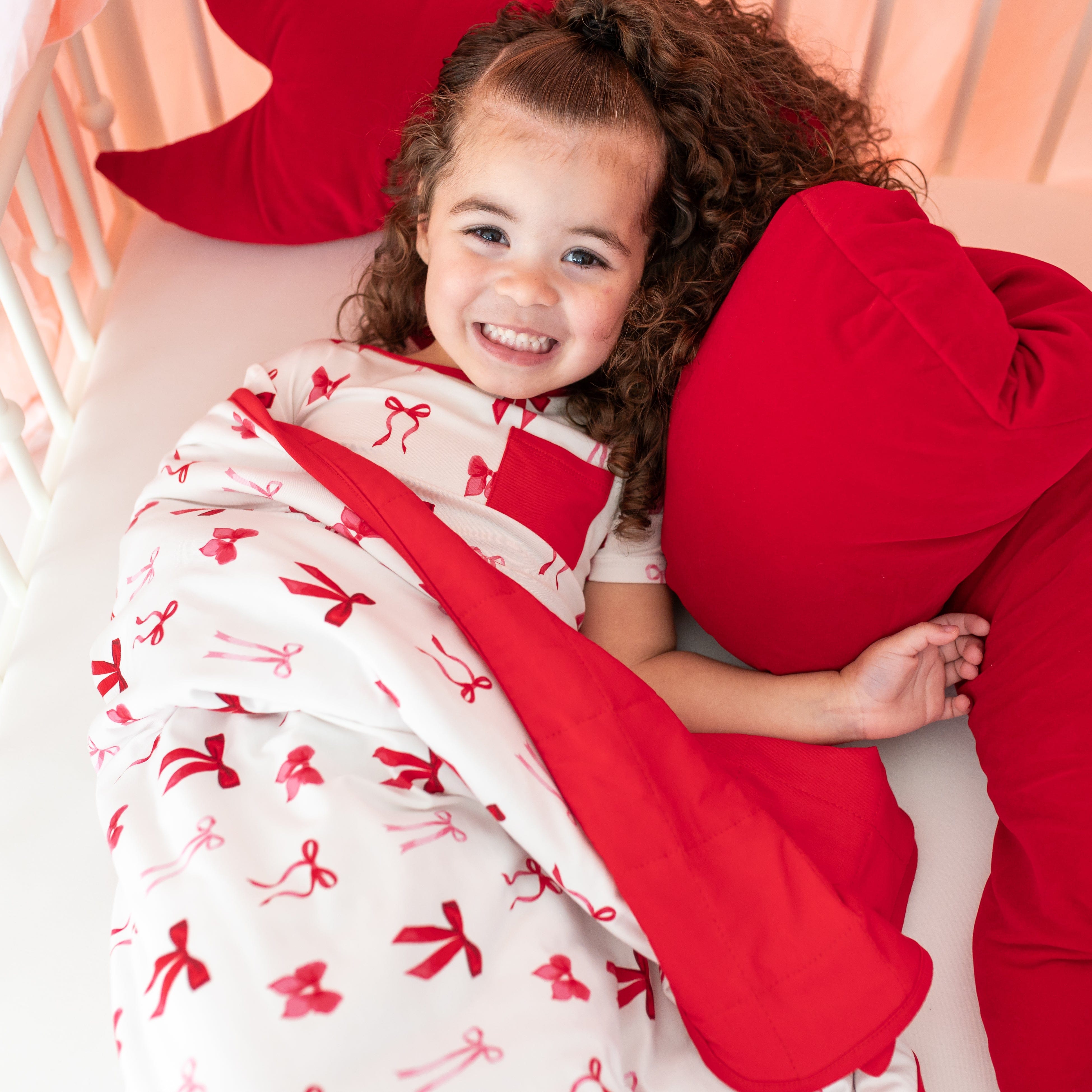 Young smiling girl laying in bed with the Toddler Blanket in Small Love Bow 1.0 on top of her