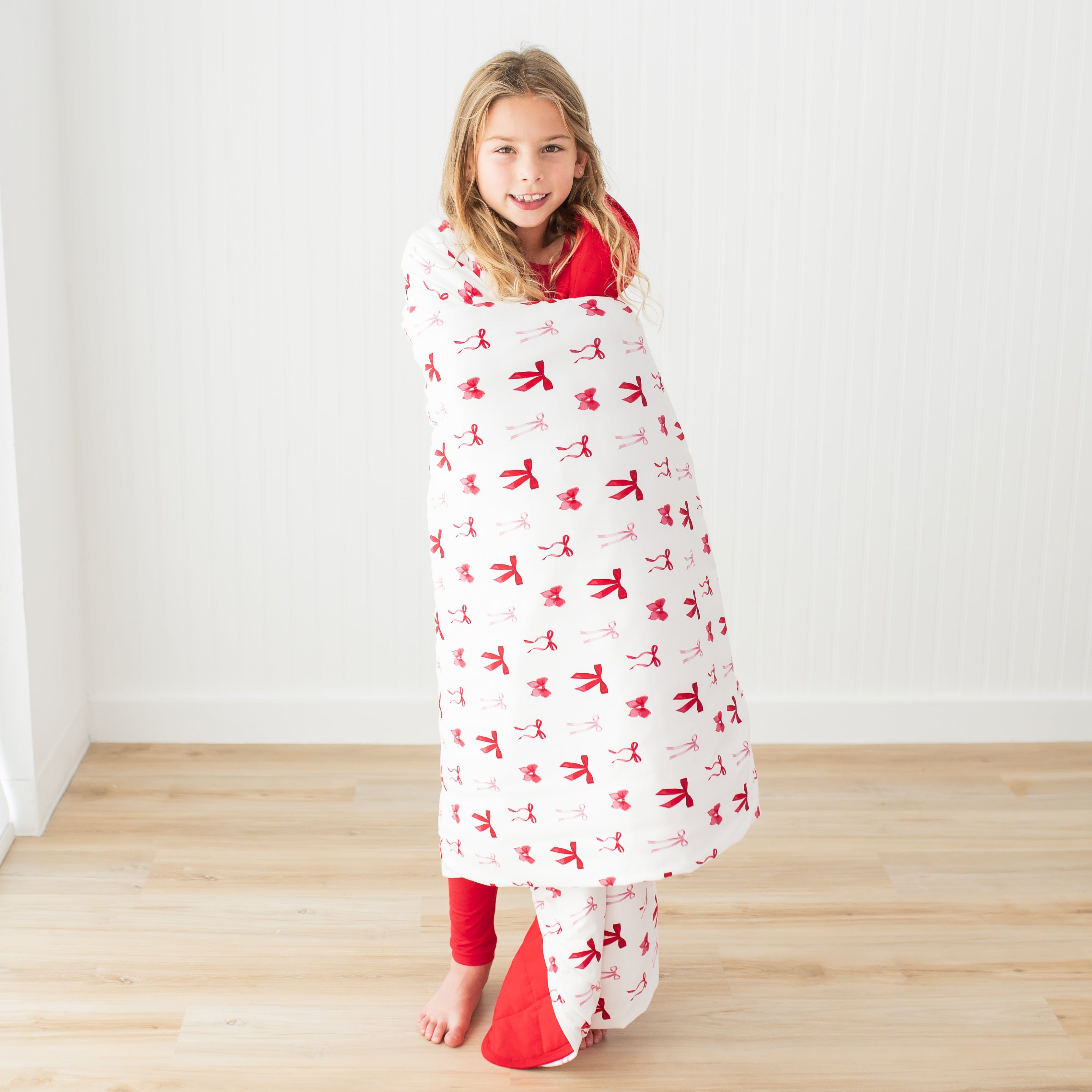 Young girl standing in front of a white paneled wall with the Youth Blanket in Small Love Bow 2.5 around her shoulders