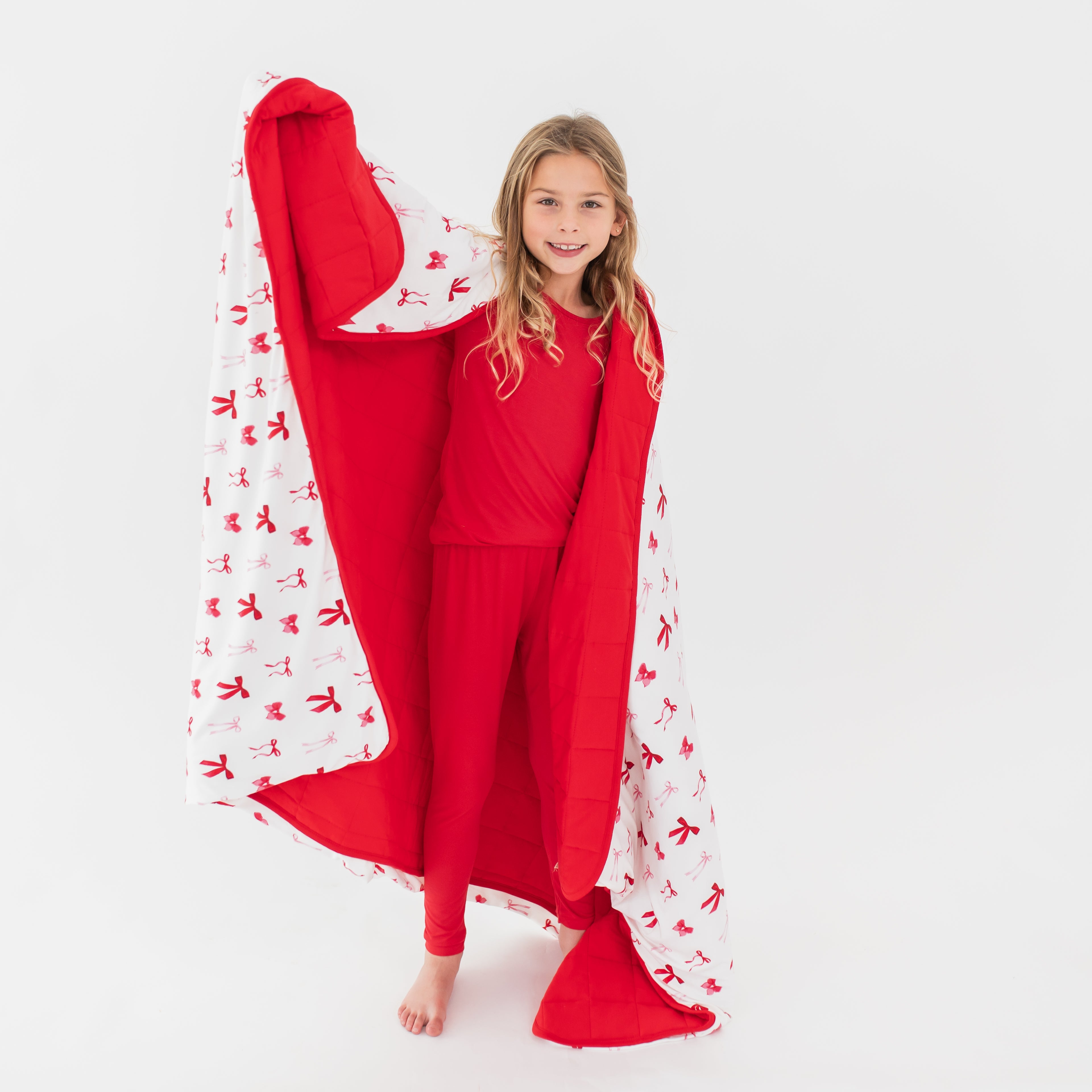 Young girl standing in front of a white background with the Youth Blanket in Small Love Bow 2.5 around her shoulders showing the red backing and printed front
