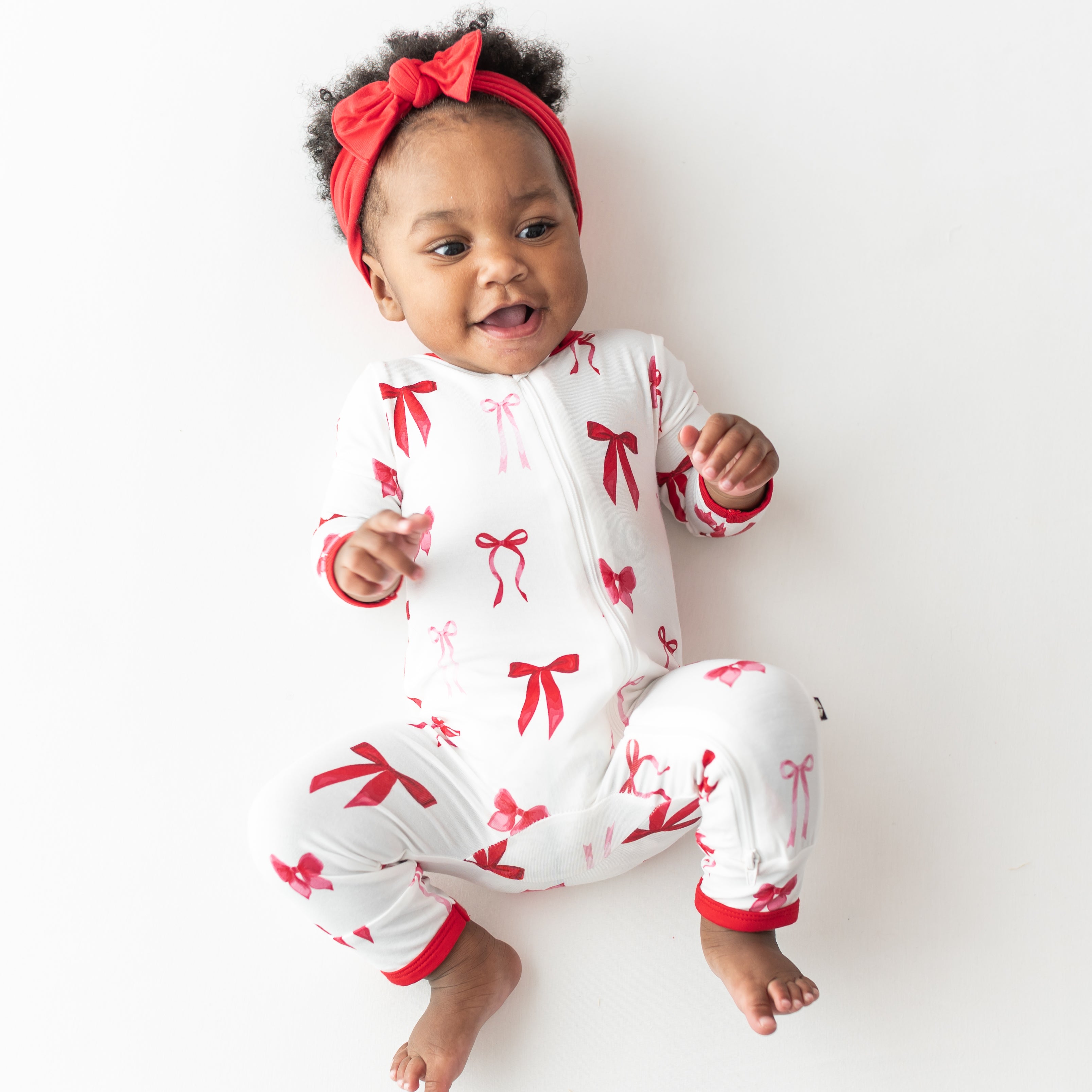 Young toddler laying on a light cream surface wearing the Zippered Romper in Small Love Bow with a red bow