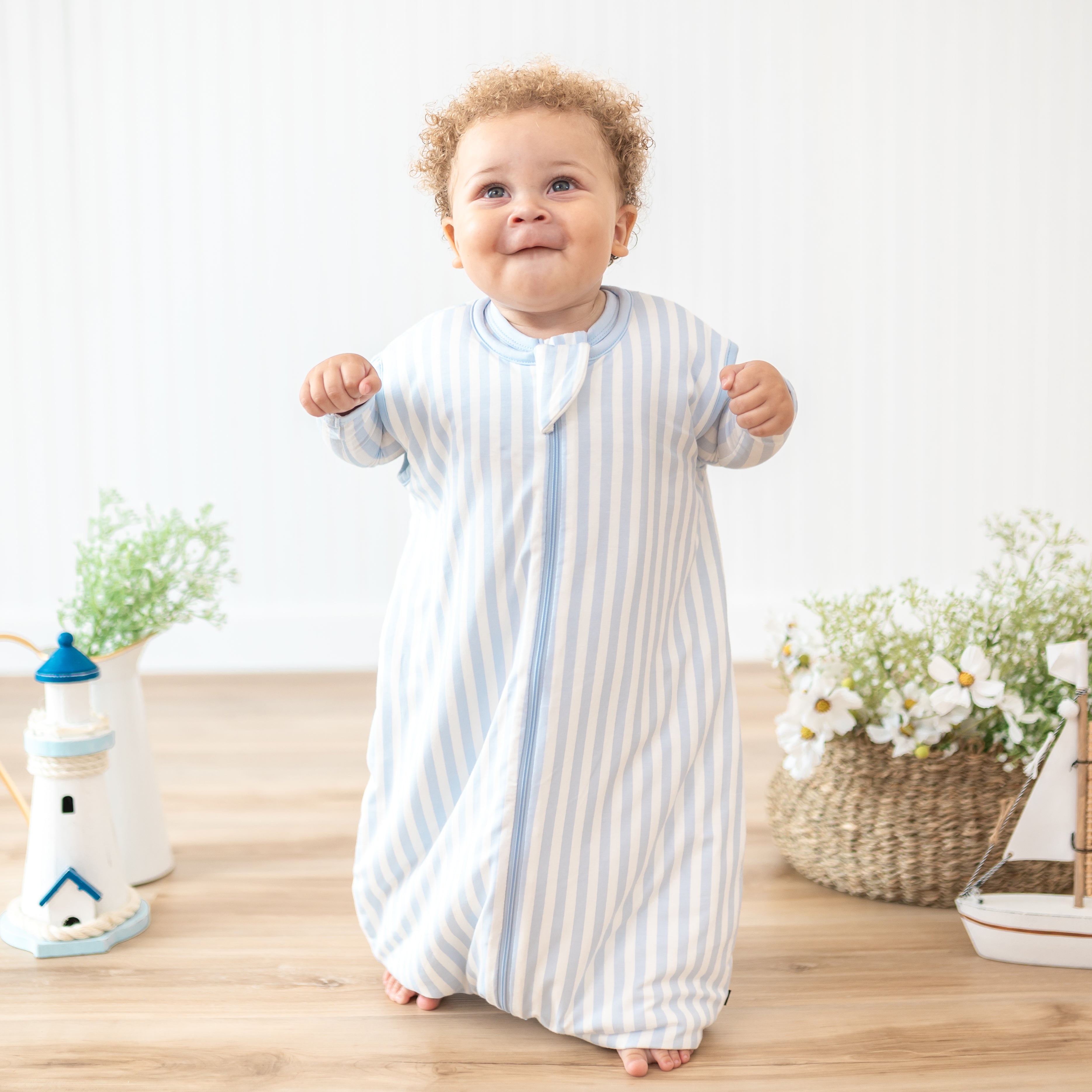 Toddler standing beside potted plants and flowers wearing the Sleep Bag Walker in Small Mist Stripe 1.0