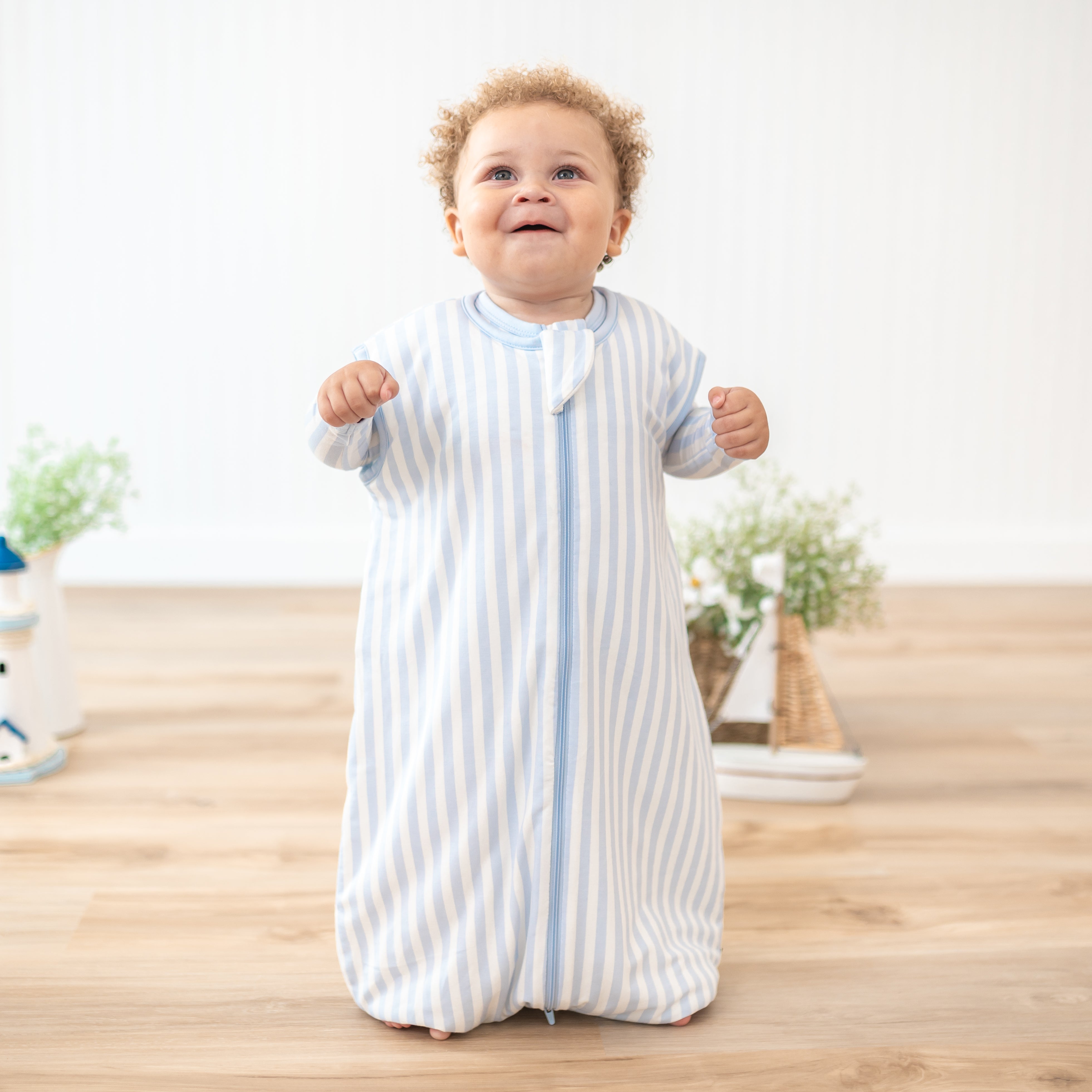 Smiling toddler standing wearing the Sleep Bag Walker in Small Mist Stripe 1.0 with matching long sleeve romper underneath