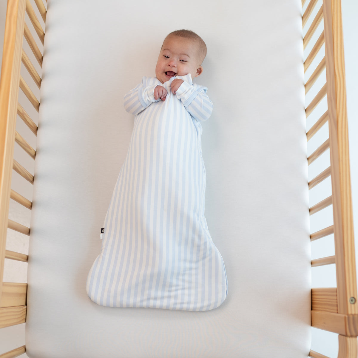 Smiling infant laying in a crib wearing the Sleep Bag in Small Mist Stripe 1.0 with matching long sleeve romper