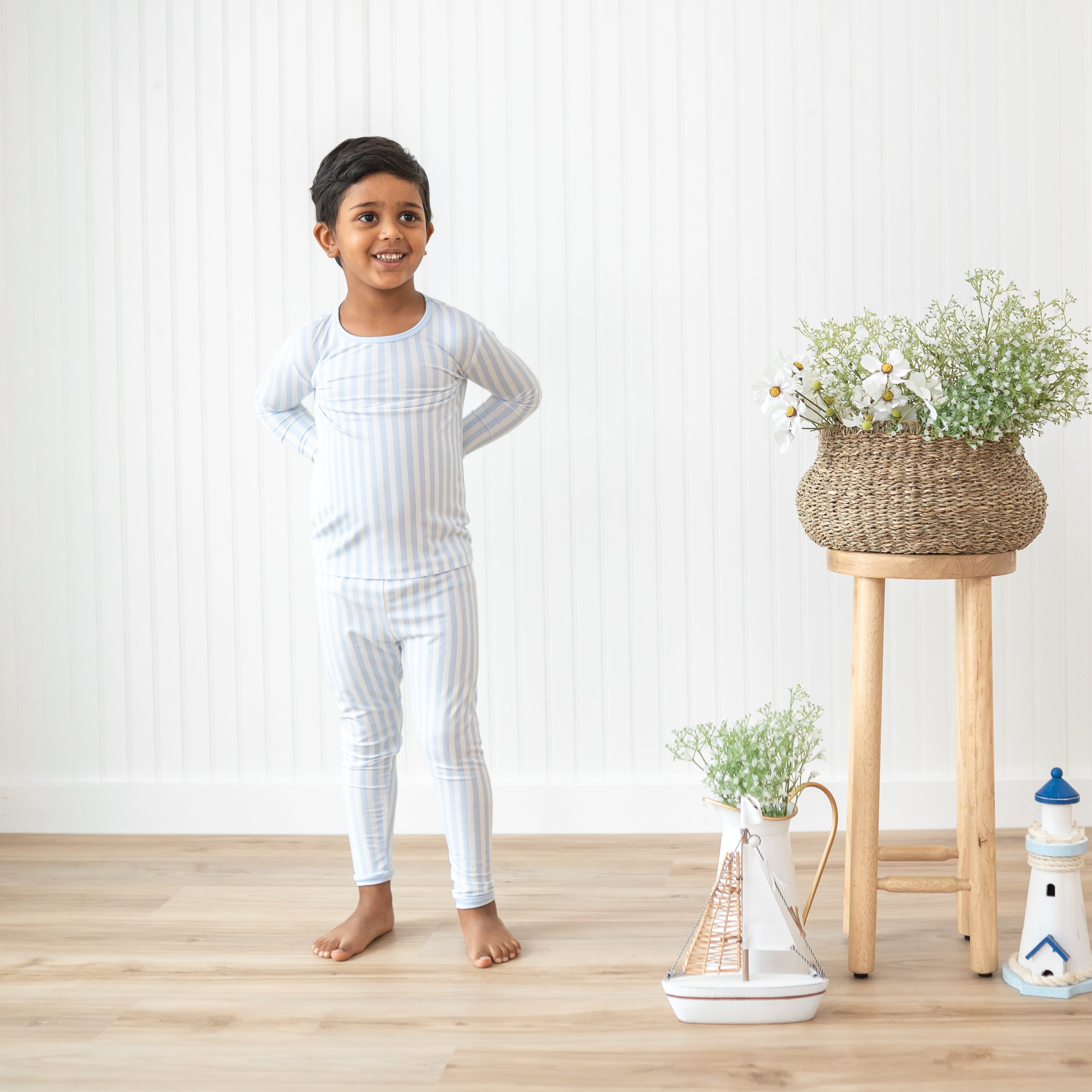 Boy modeling the Long Sleeve Pajamas in Small Mist Stripe beside potted flowers and a nautical themed toys