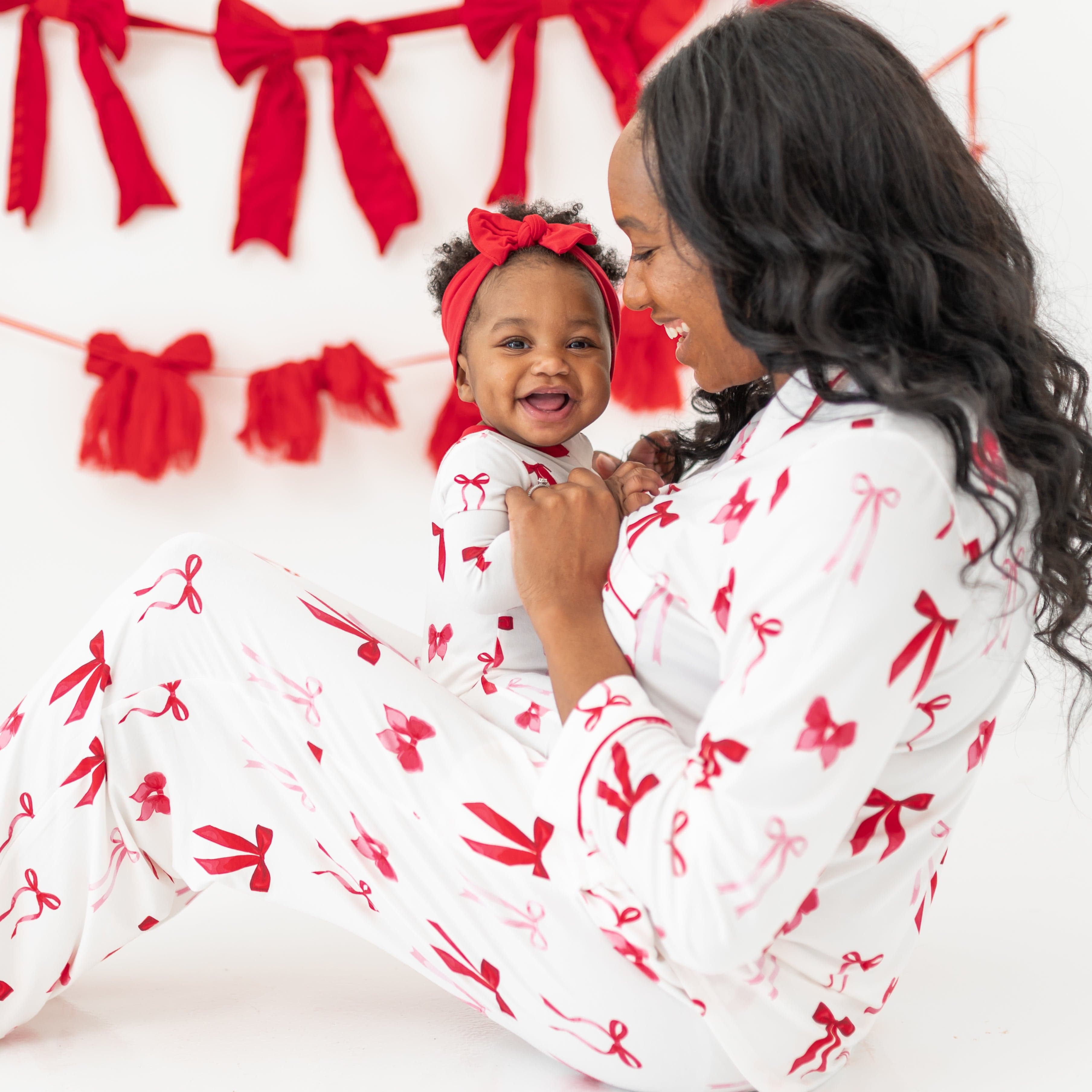 Mother sitting on the floor wearing the Women's Pajama Set in Big love bow holding her young toddler who is matching in a Zippered Romper in Small Love Bow