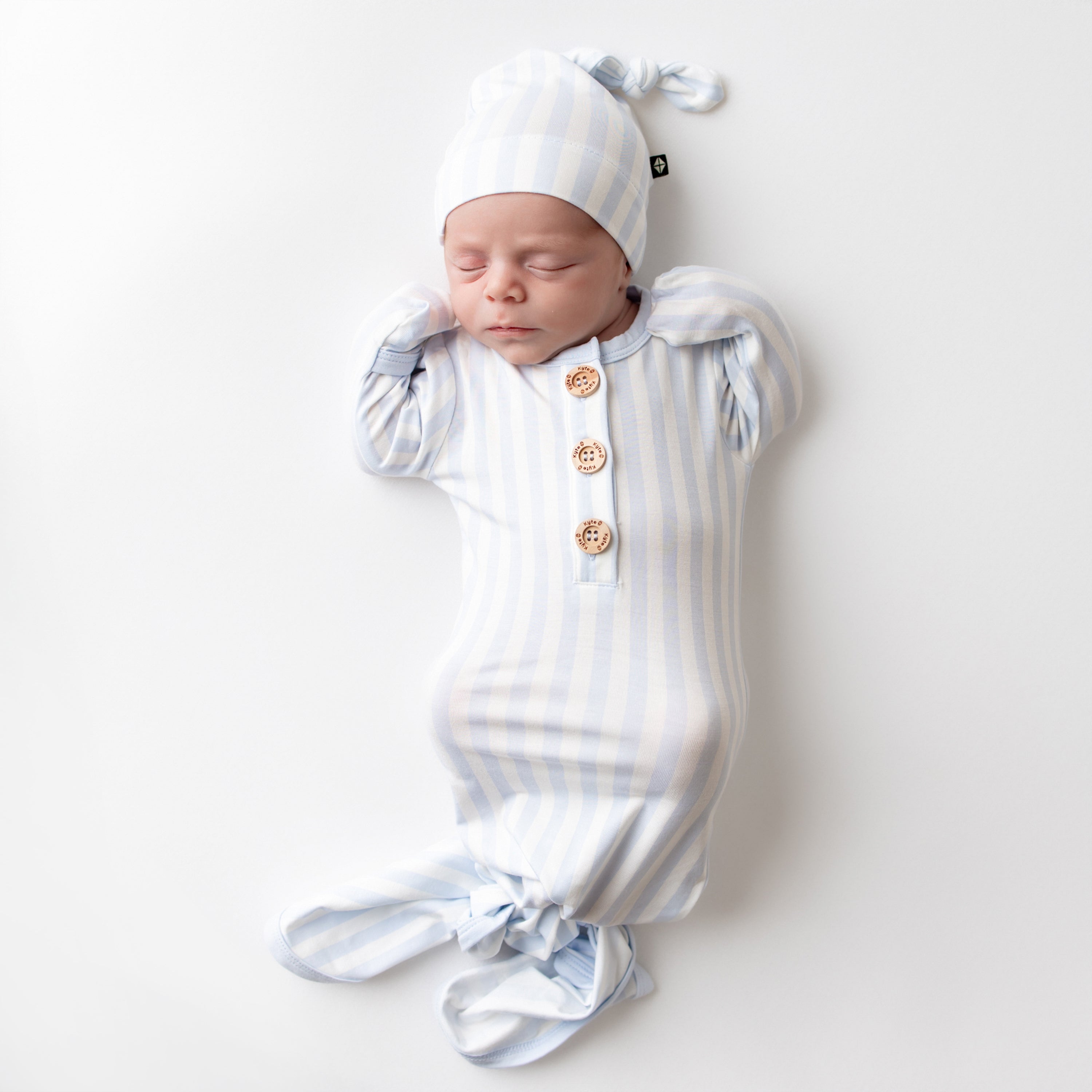 Sleeping newborn laying down on a light neutral surface wearing the Knotted Gown with Hat Set in Small Mist Stripe