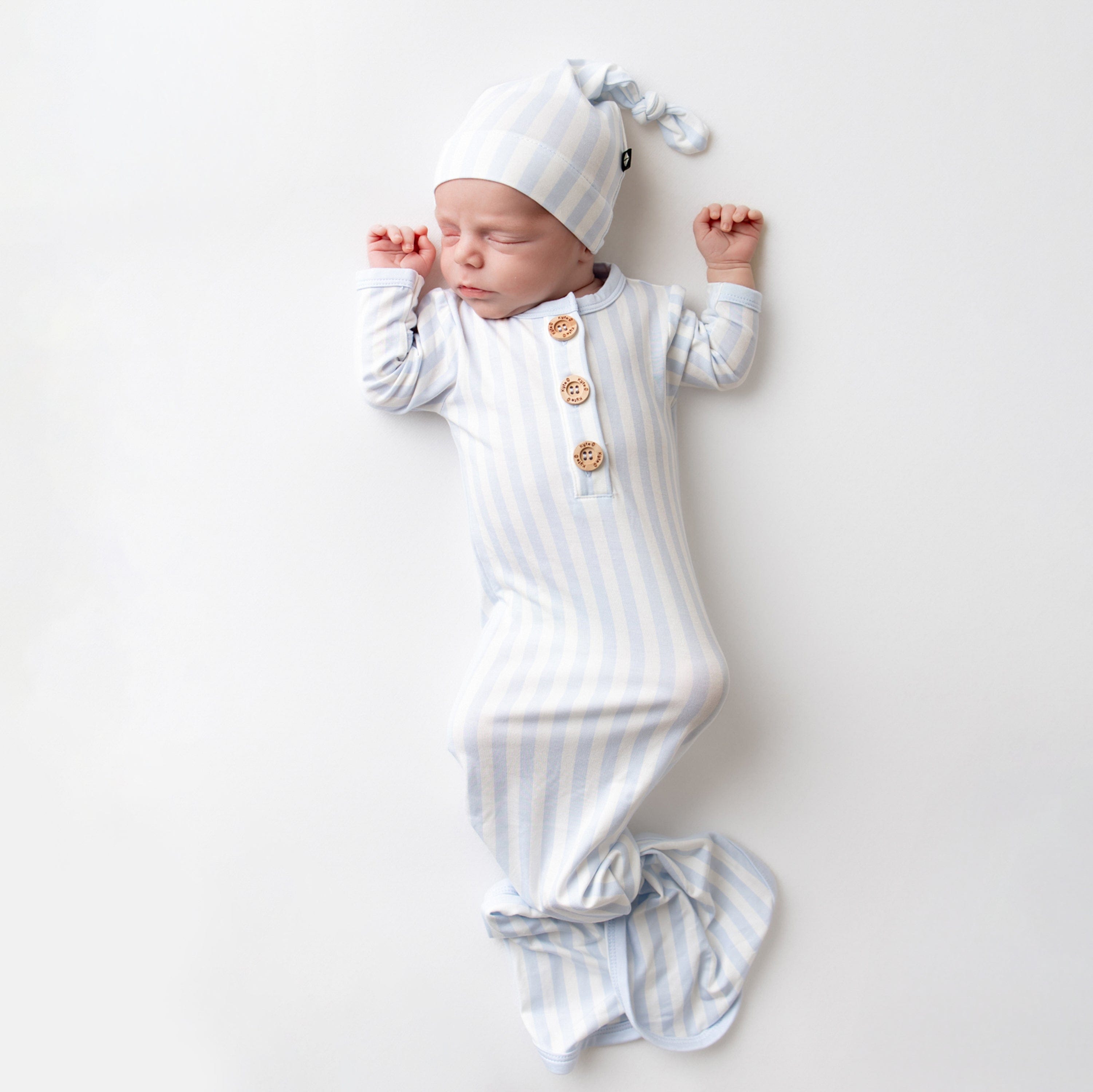 Sleeping newborn laying on a light neutral surface wearing the Knotted Gown with Hat Set in Small Mist Stripe