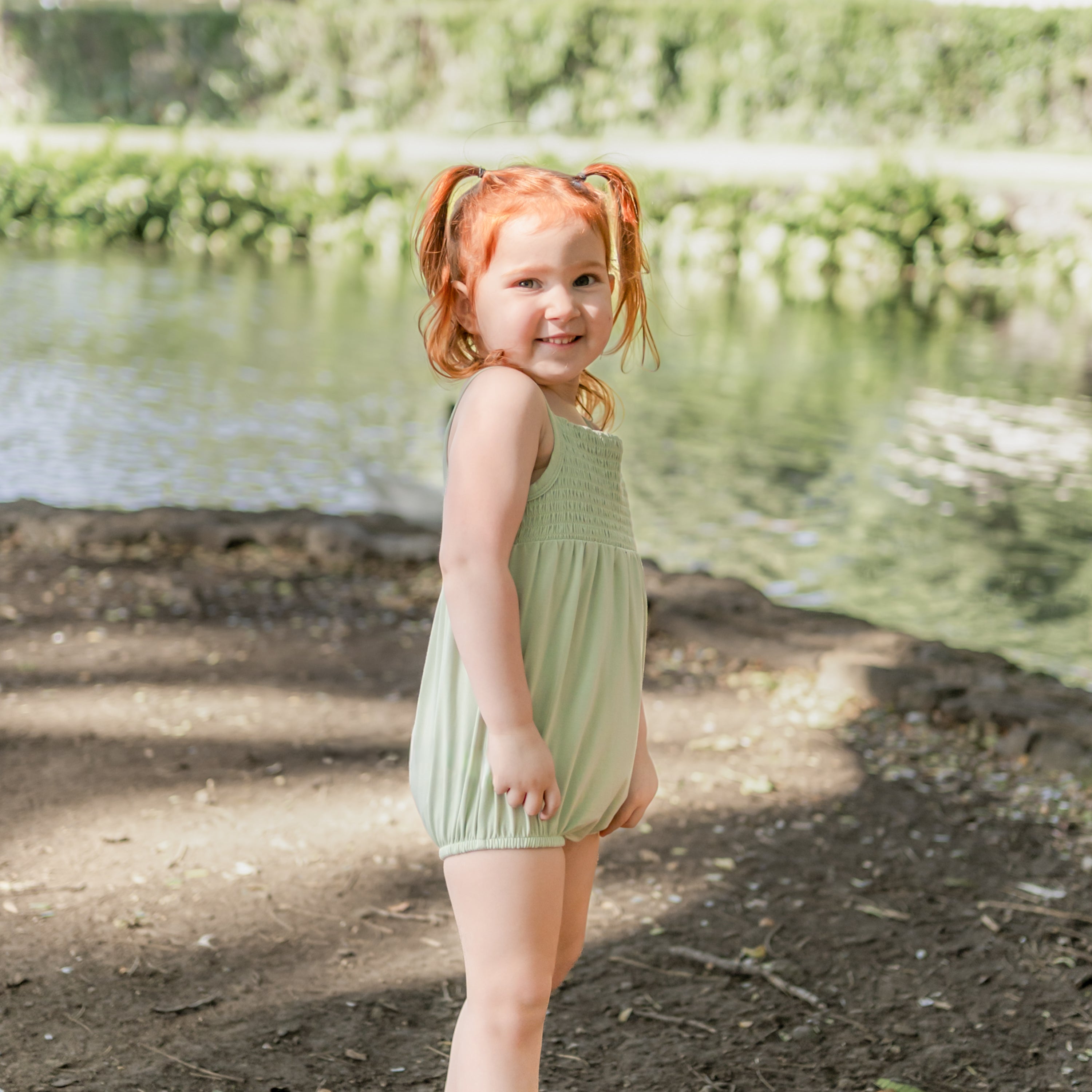 Young girl standing by the water edge wearing the Smocked Bubble Romper in Basil