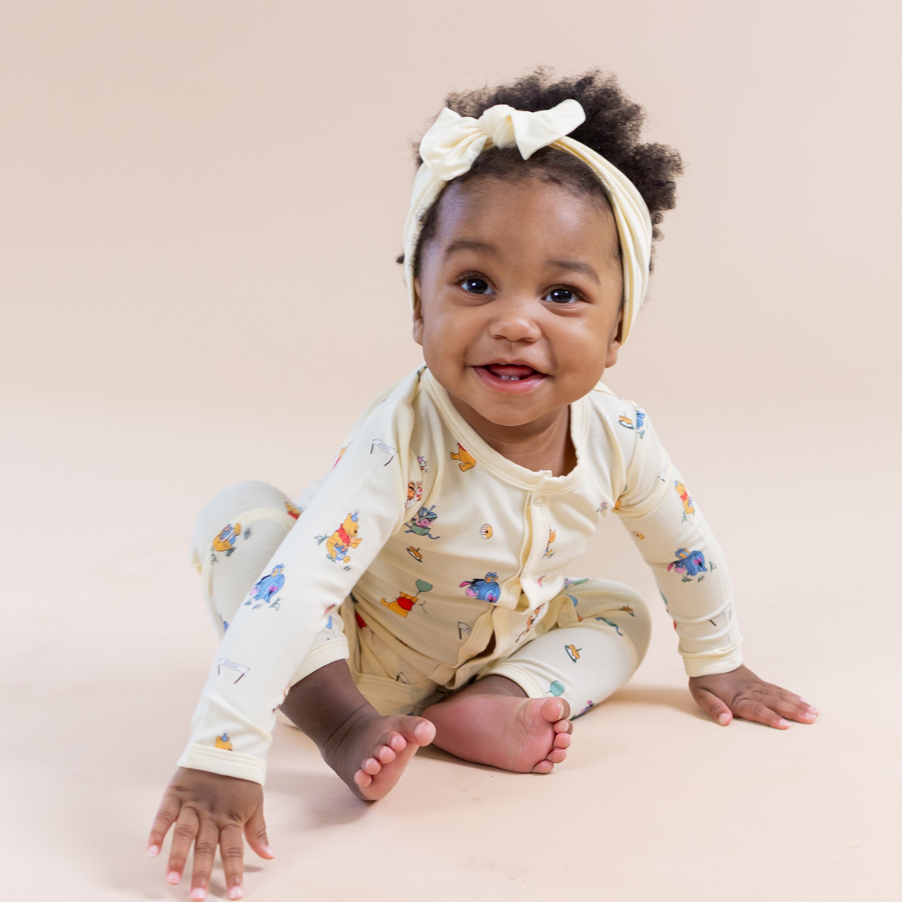 Smiling toddler sitting on the floor wearing the Romper in Winnie the Pooh Celebration with a light yellow bow