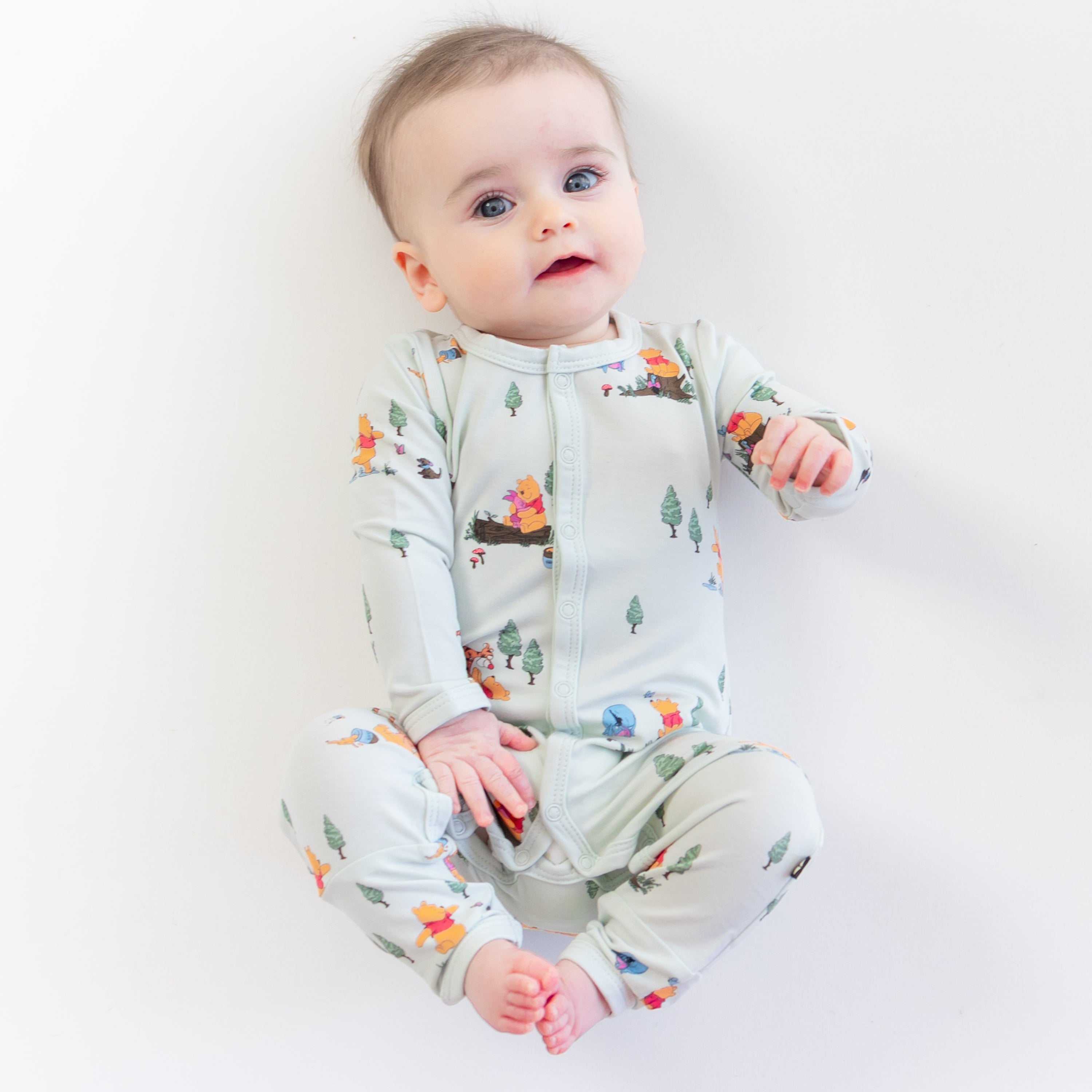 Young infant laying on a light neutral surface wearing the Romper in Winnie the Pooh Hundred Acre Wood